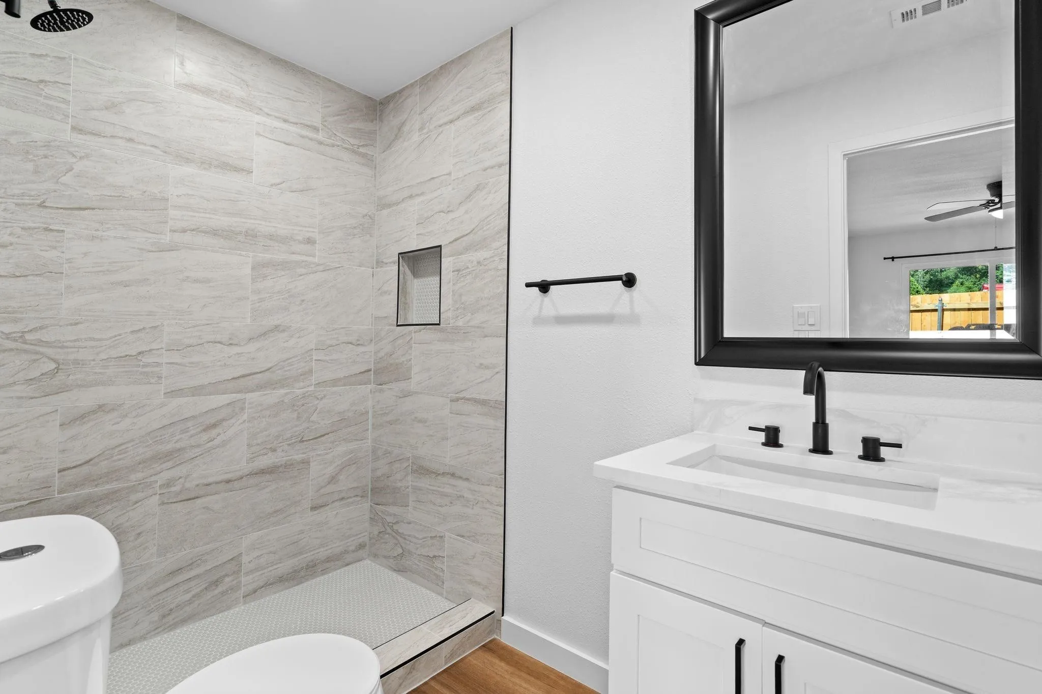 Bathroom featuring a vanity with a stone-surface countertop, matte black widespread faucet, framed mirror, and walk-in shower with stone-style wall tile and a built-in recessed niche