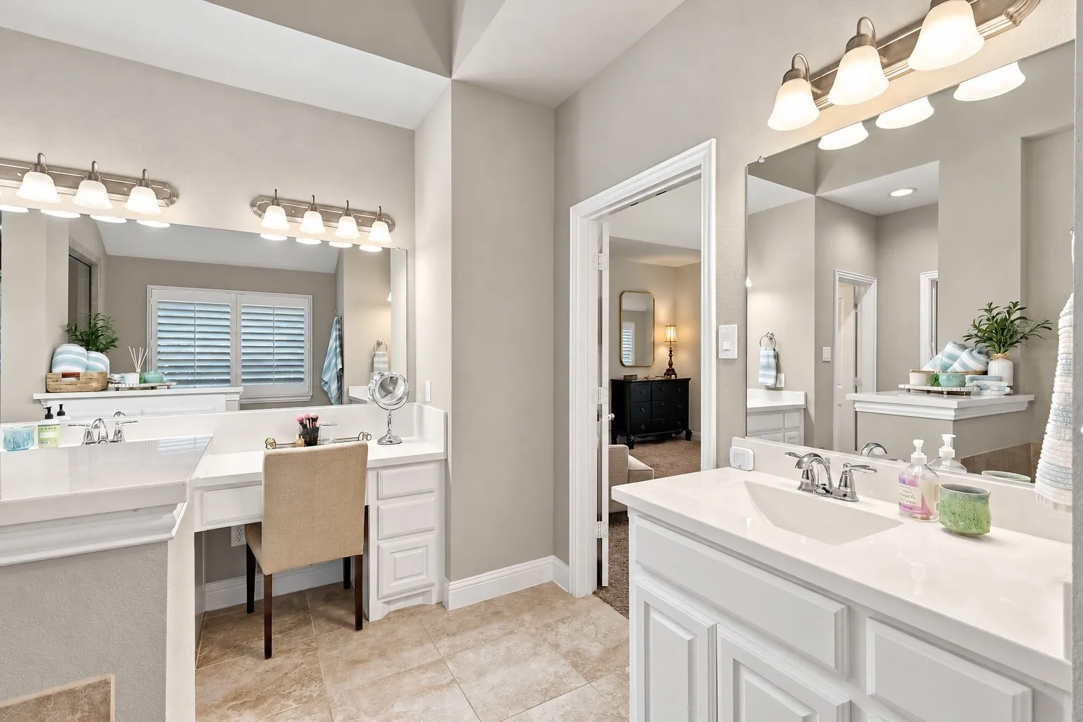 Dual vanity featuring white cabinetry, solid-surface countertops, integrated sinks, framed mirrors, and multi-light wall fixtures, complemented by a built-in makeup vanity and tiled flooring