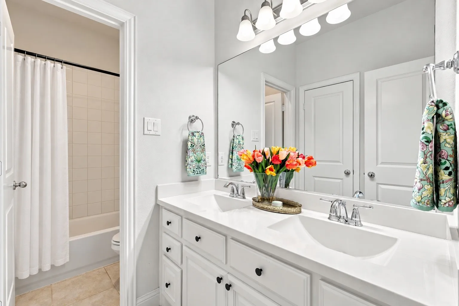 Dual-sink vanity with white cabinetry, light-toned stone countertop, chrome hardware, and a large-format vanity mirror with integrated light fixture