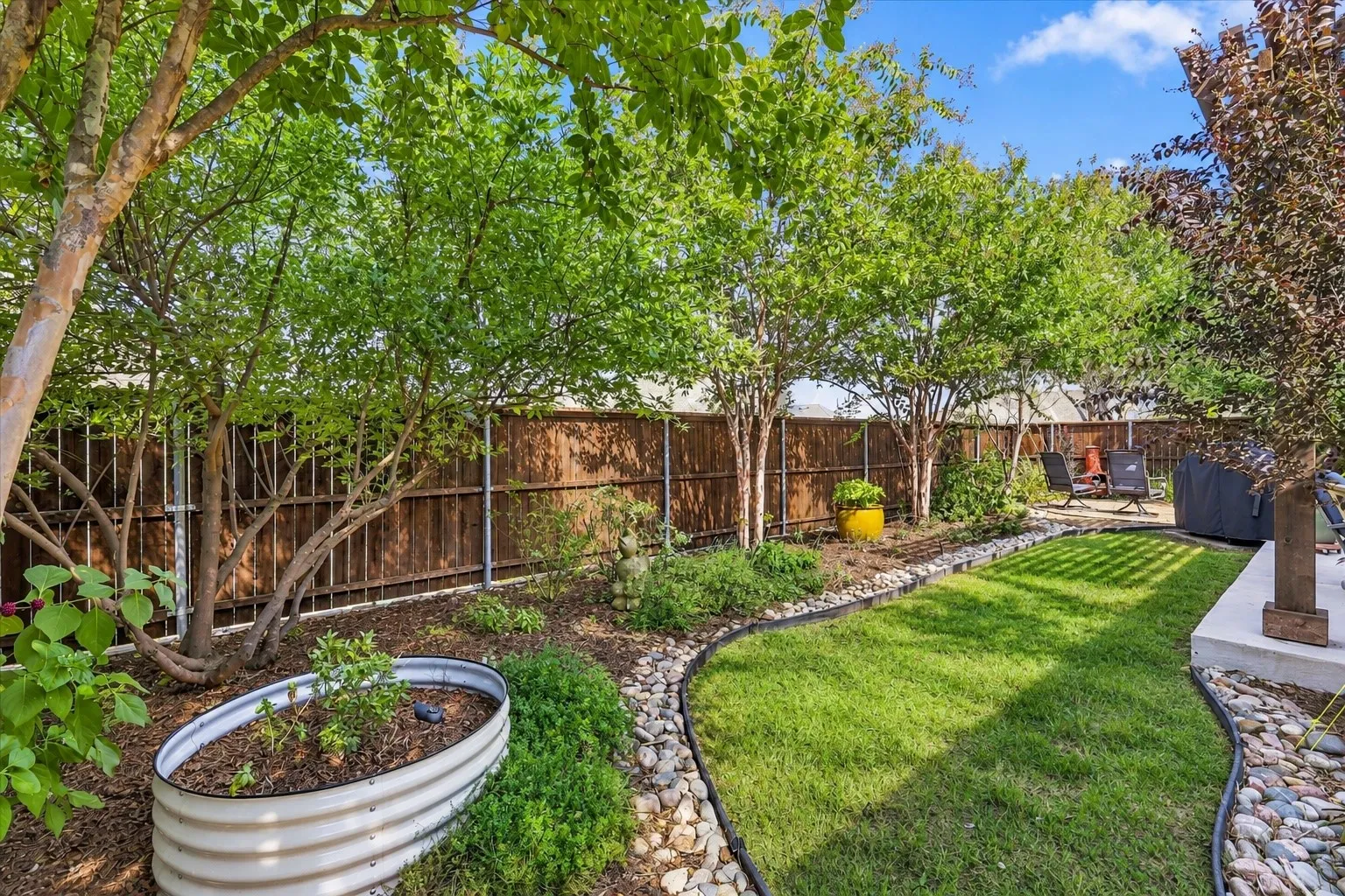 Backyard featuring a manicured lawn, stone border edging, mature trees, metal raised garden bed, and wood-panel privacy fencing