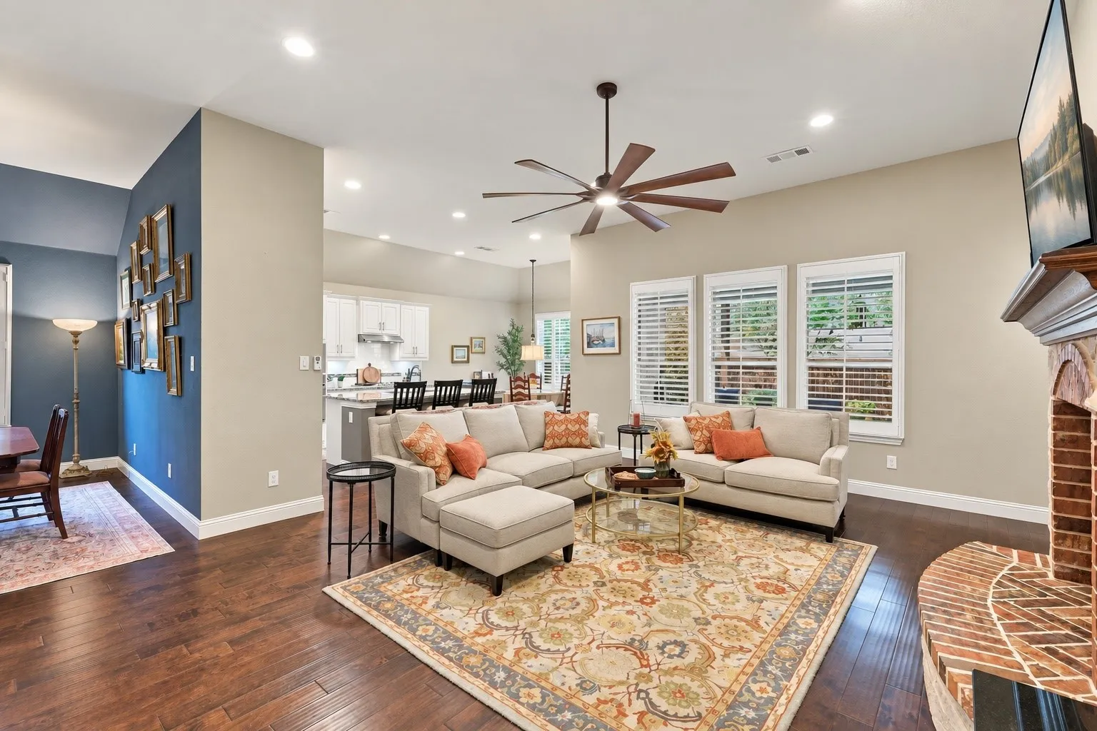 Living area featuring wood-finish flooring, a vaulted ceiling with a multi-blade fan, custom window shutters, and a brick-accented fireplace hearth