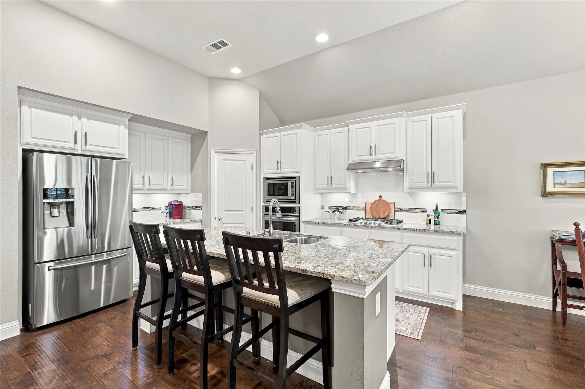 Kitchen featuring white shaker-style cabinetry, stone countertops, stainless steel appliances, a center island with sink, and dark wood-finish flooring