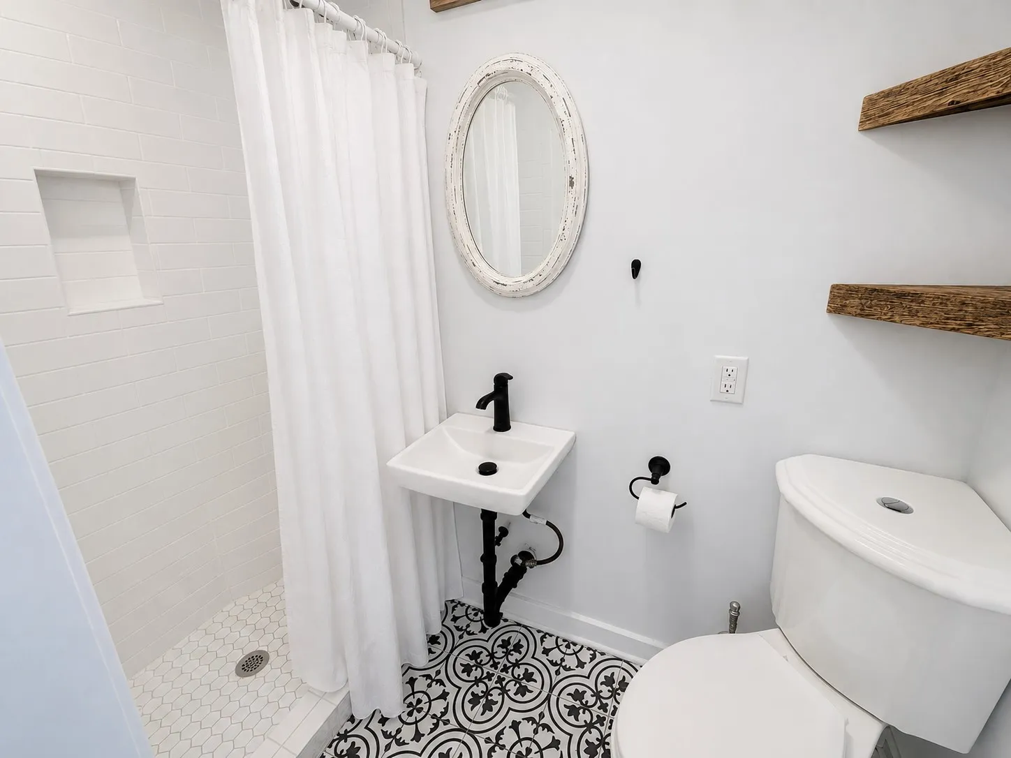 Bathroom featuring patterned floor tile, wall-mounted sink with matte black faucet, shower with integrated wall niche, and natural wood-finish floating shelving