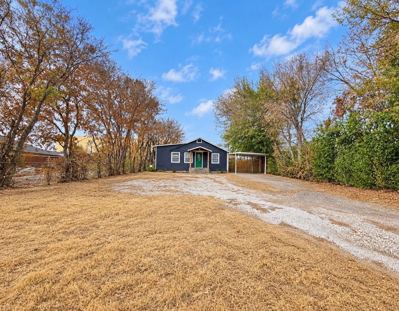 Single-story exterior featuring dark lap siding, a covered carport, a gravel driveway, and mature perimeter trees