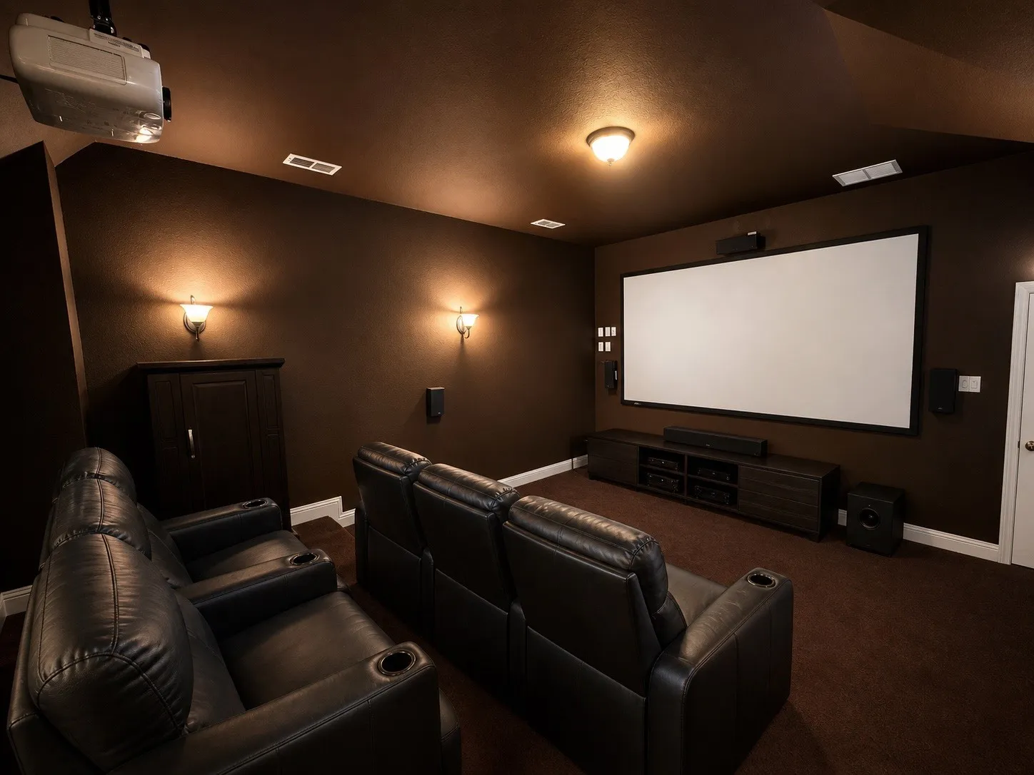 Dedicated home theater featuring a large projection screen, dark-toned wall treatment, recessed ceiling lighting, and wall sconces