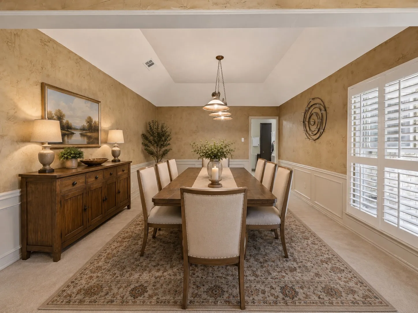 Tray ceiling with a suspended light fixture, wainscoting, textured wall finish, and window with plantation shutters