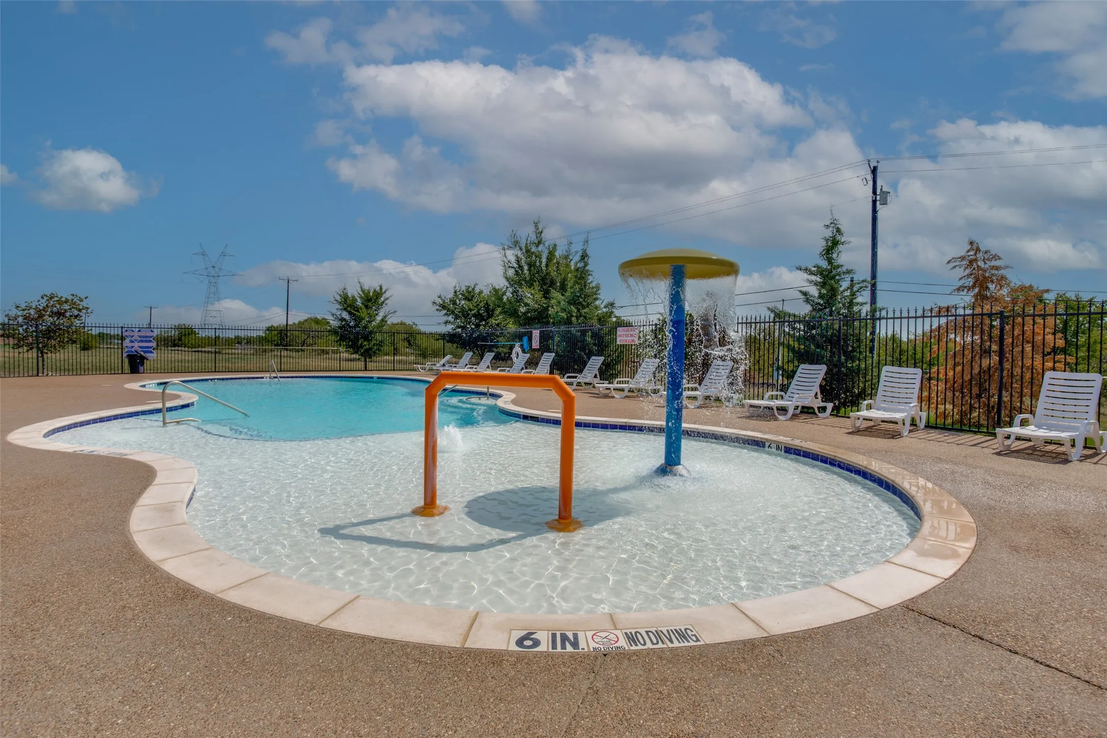 Swimming pool featuring a zero-entry design, water play structure, and concrete decking enclosed by perimeter fencing