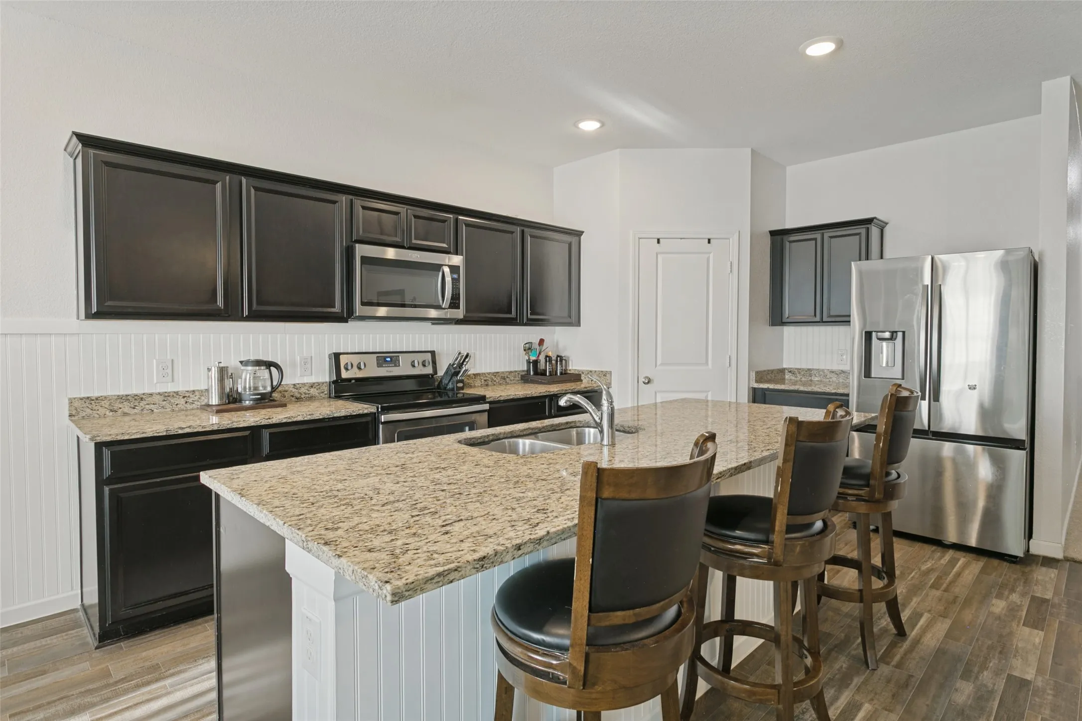 Kitchen featuring dark cabinetry, stone-surface countertops, central island with stainless steel sink, beadboard backsplash, and wood-finish flooring