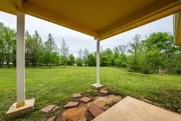 Covered concrete patio featuring structural support columns, flagstone accents, and a grass lawn with perimeter trees