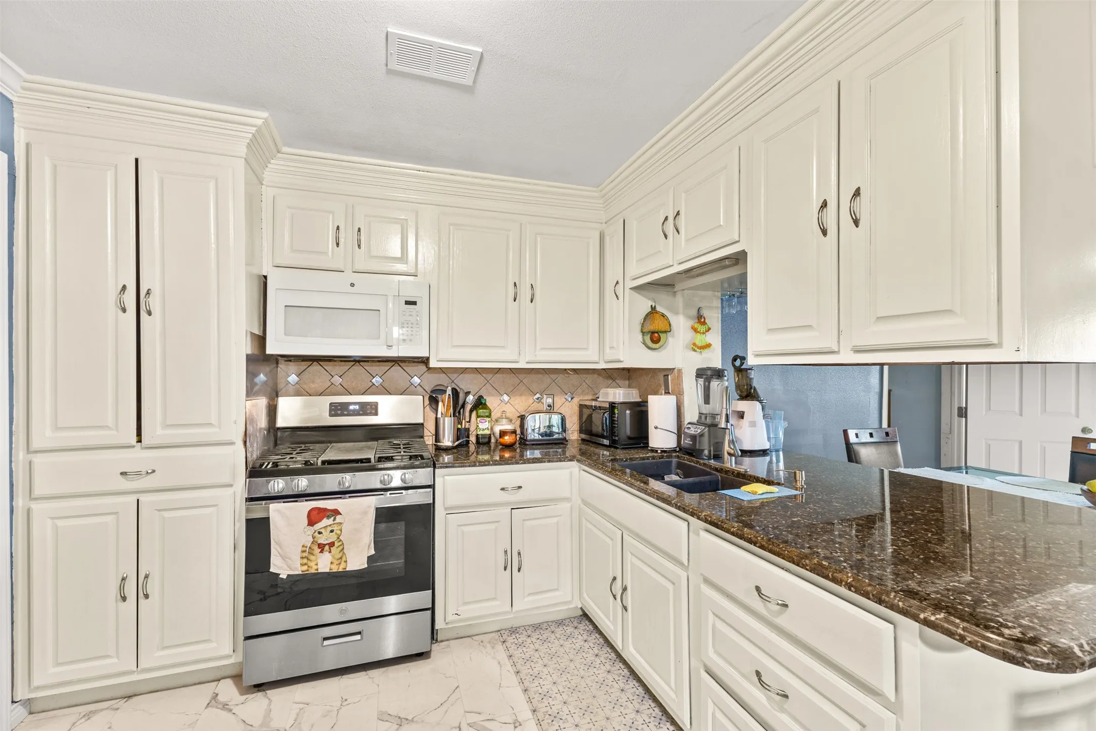 Kitchen featuring off-white cabinetry with crown molding, dark stone-finish countertops, stainless steel gas range, over-the-range microwave, and stone tile flooring