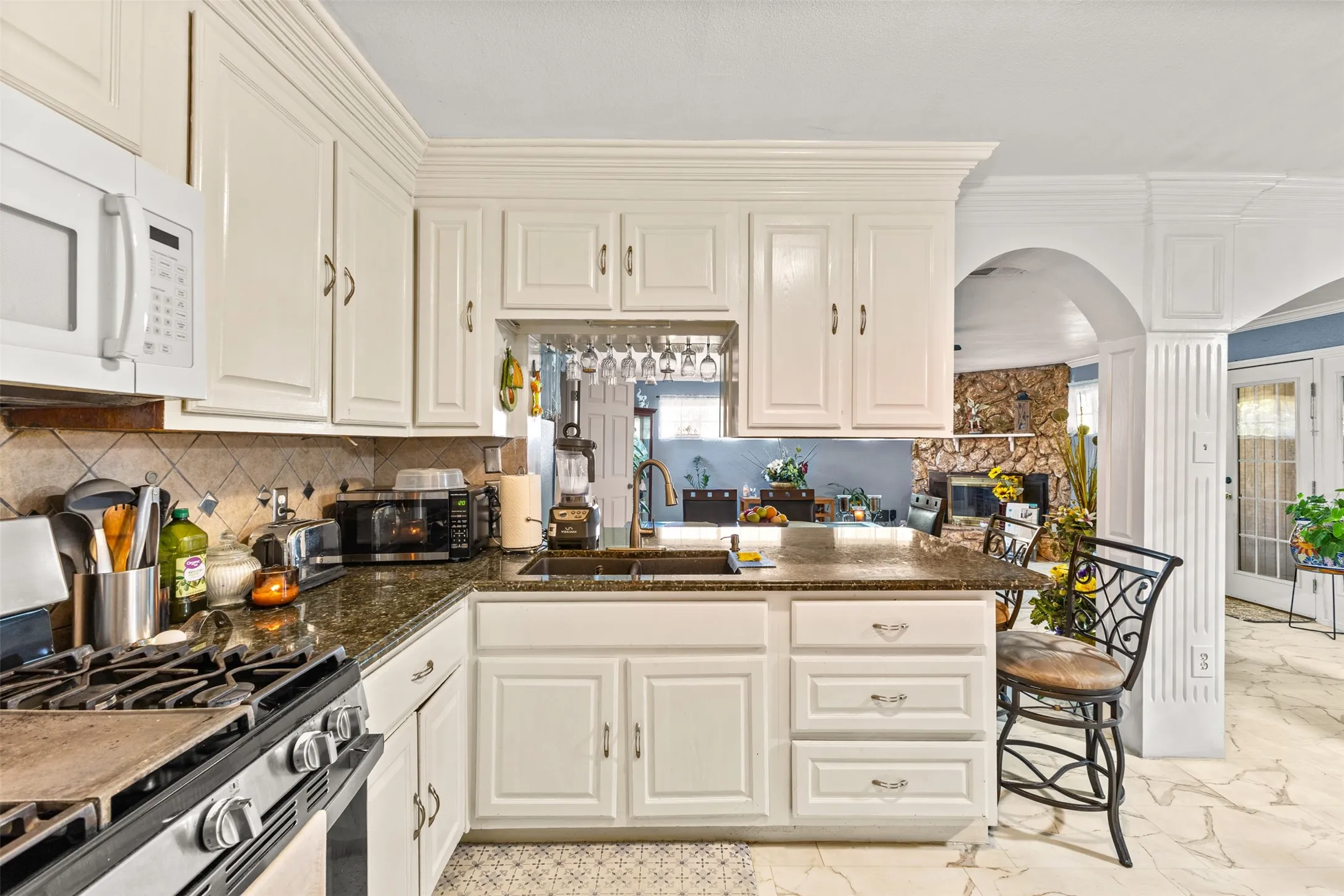 Kitchen featuring white cabinetry with crown molding, stone-finish countertops, stainless steel undermount sink with pull-down faucet, tile backsplash, and stone-finish flooring