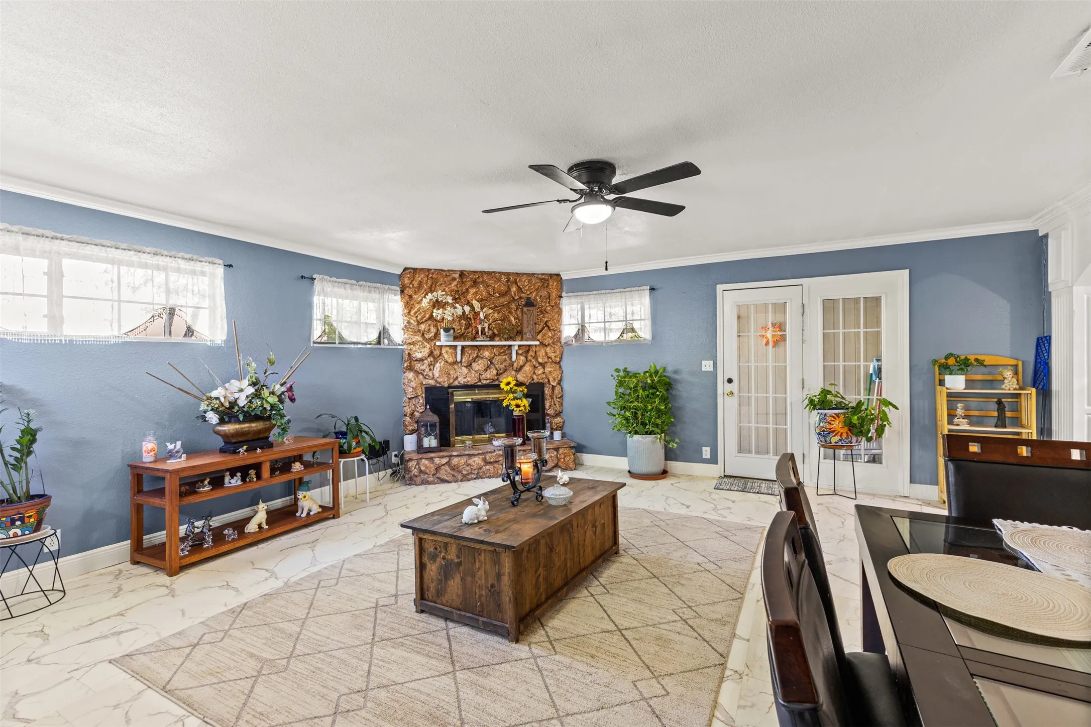 Living area featuring natural stone fireplace surround, light-toned patterned tile flooring, ceiling fan, and multi-light glass pane exterior door