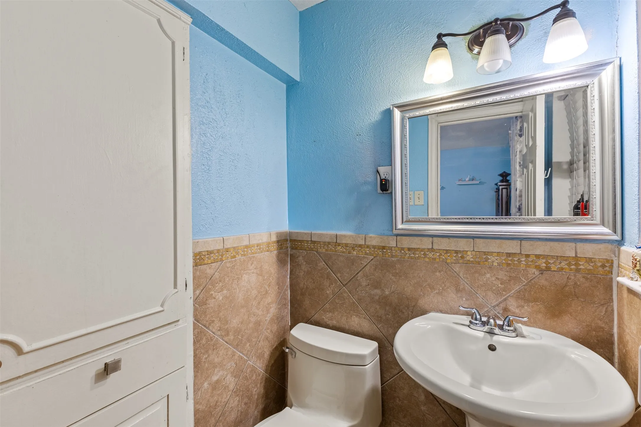 Bathroom featuring a pedestal sink with a chrome faucet, large-format diamond-patterned wall tile with decorative accent border, built-in floor-to-ceiling cabinetry, and a framed vanity mirror with tri-light fixture