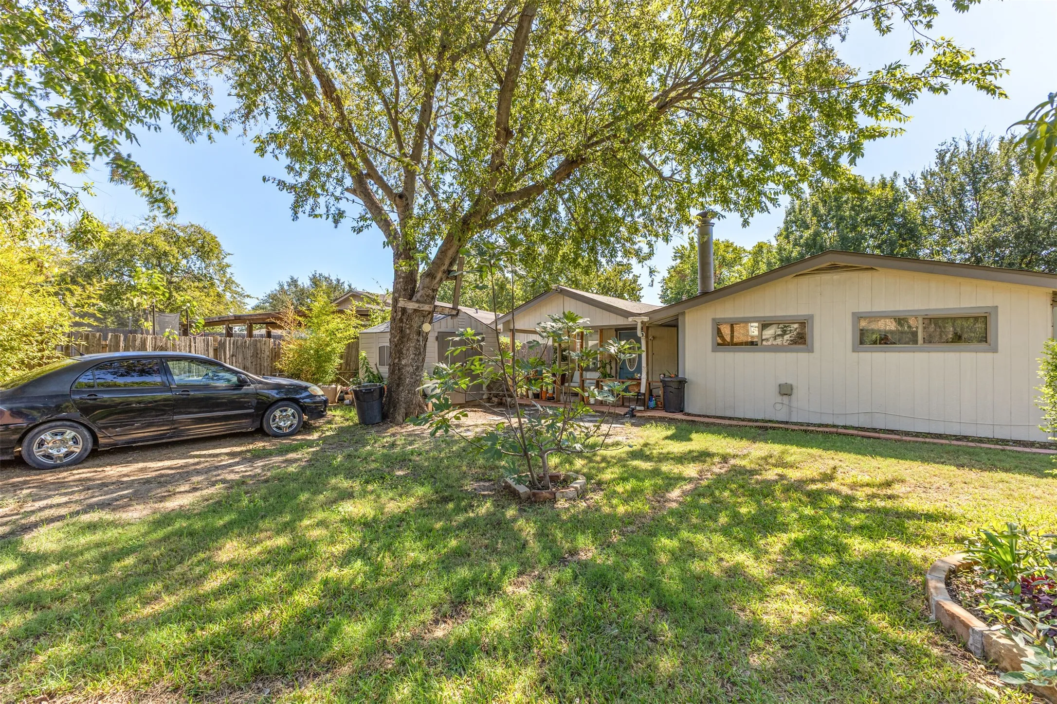 Backyard area featuring mature trees, a dedicated gravel parking space, a detached storage structure, and brick-edged garden beds