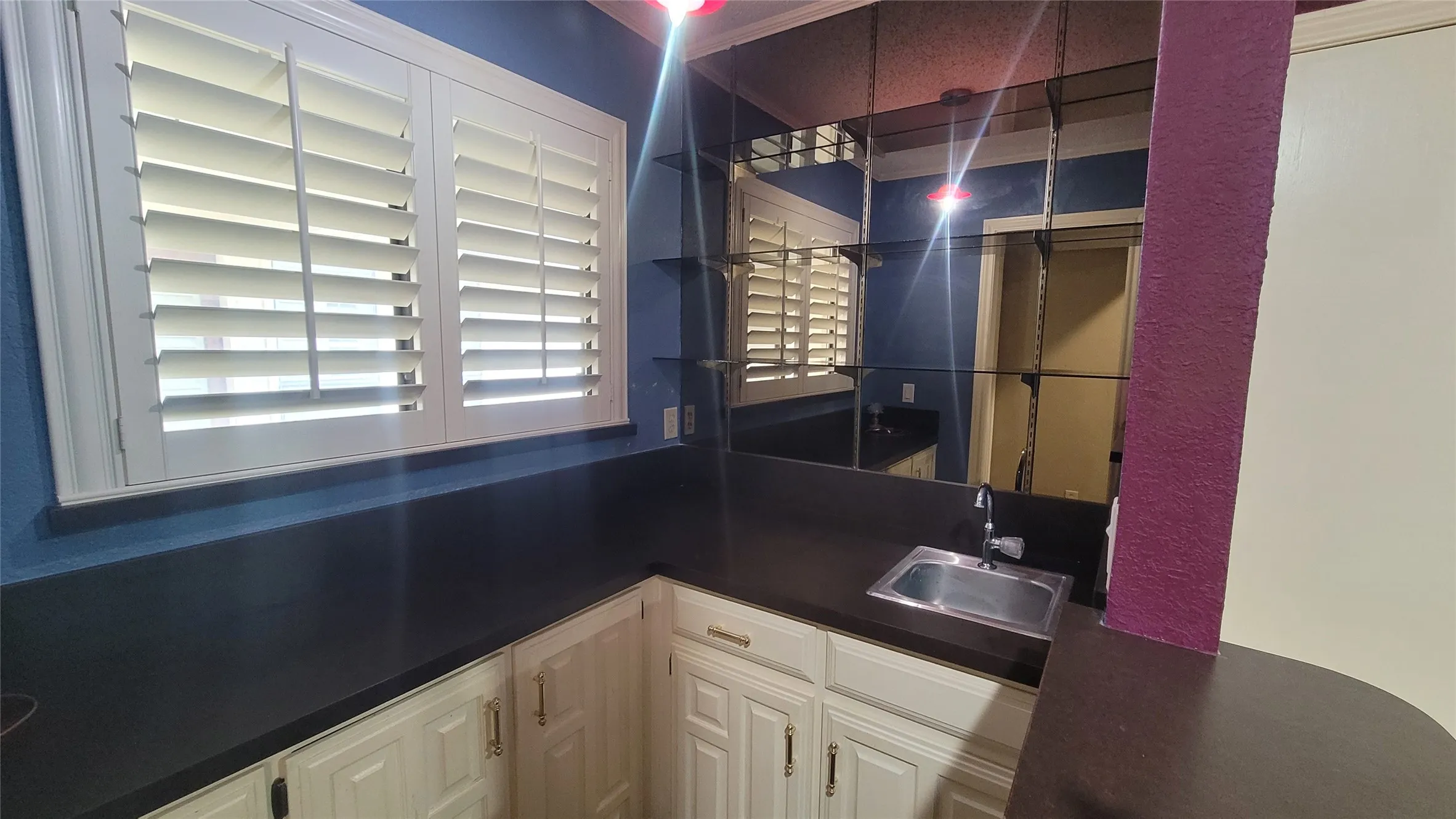 Wet bar featuring custom white cabinetry, dark solid-surface countertops, integrated stainless steel sink, mirrored backsplash, and louvered window shutters