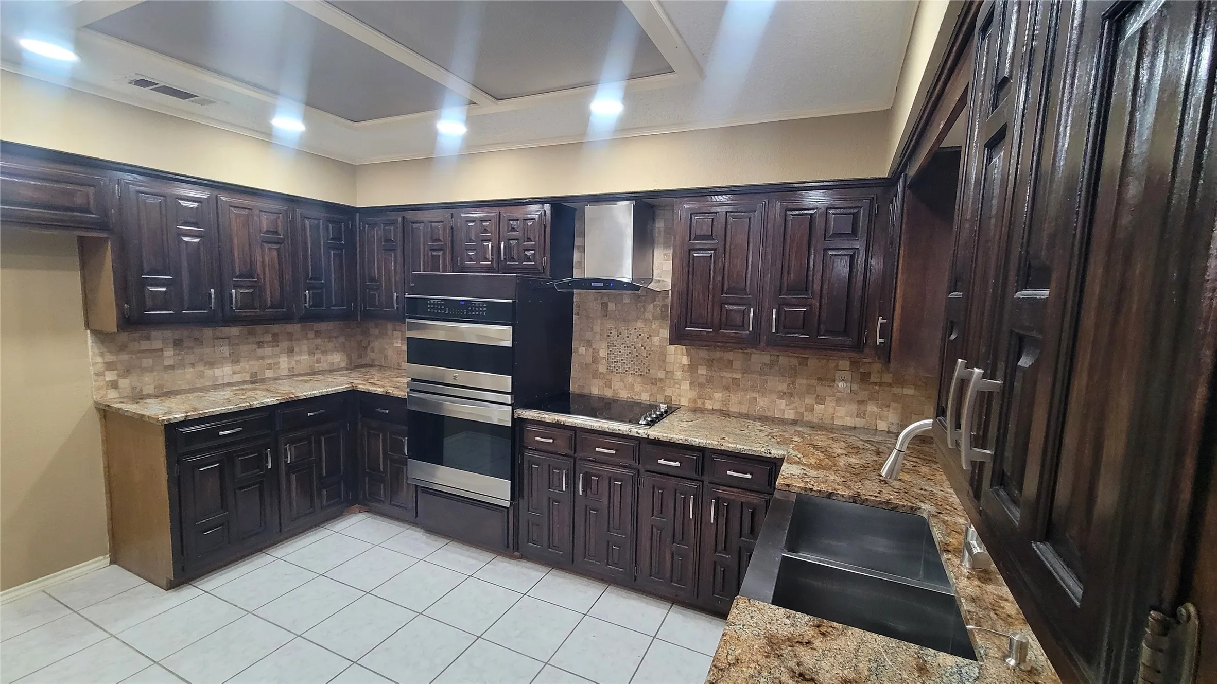 Kitchen featuring dark wood-finish cabinetry, natural stone countertops, a mosaic tile backsplash, stainless steel built-in double oven, and a recessed ceiling panel