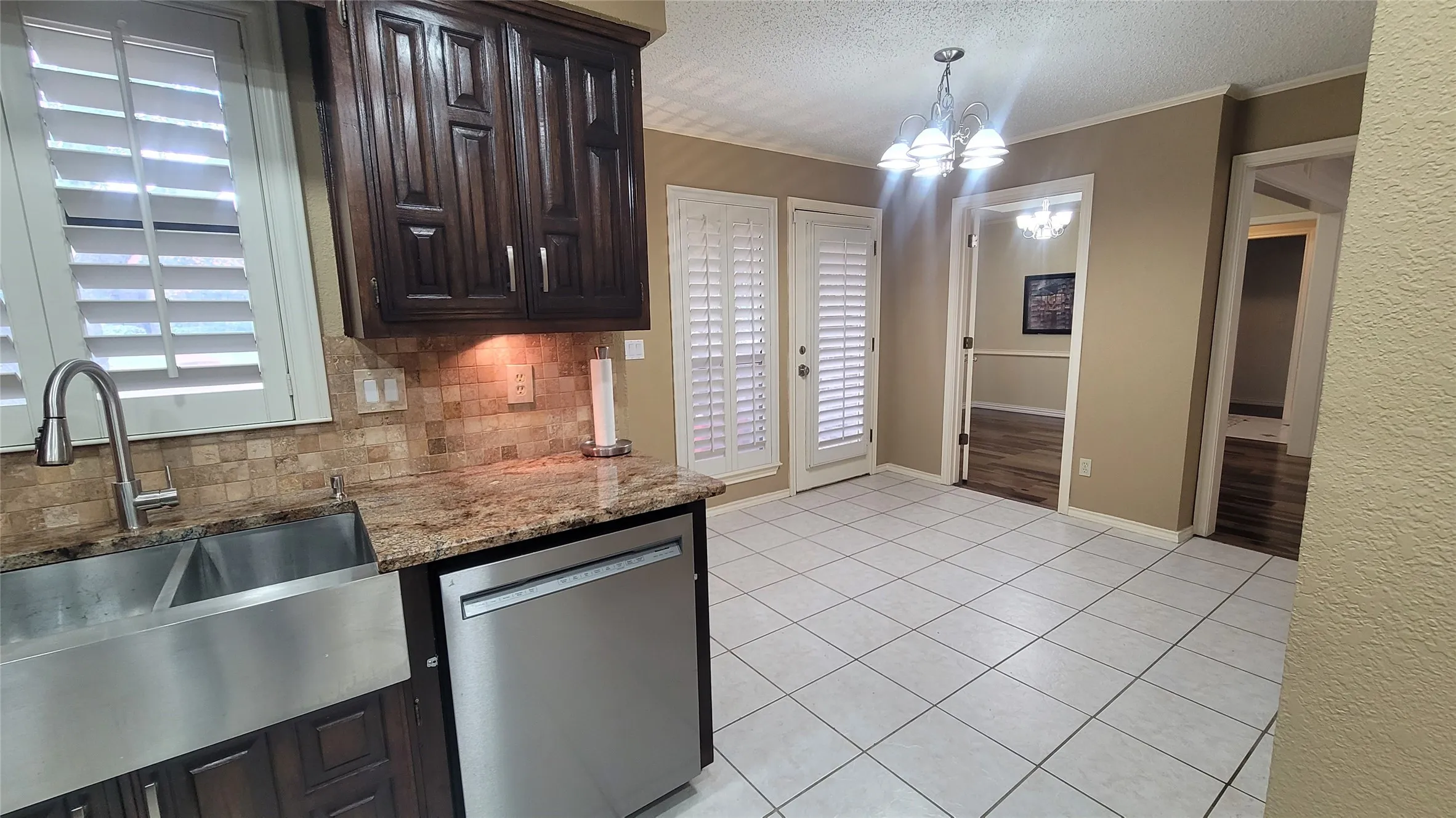 Kitchen featuring dark wood cabinetry, stainless steel apron-front sink, stone-surface countertops, tile backsplash, and plantation shutters