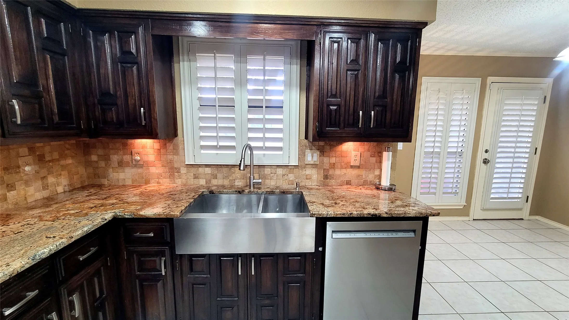 Kitchen featuring dark wood-finish cabinetry, natural stone countertops, tile backsplash, stainless steel farmhouse sink, and plantation shutters