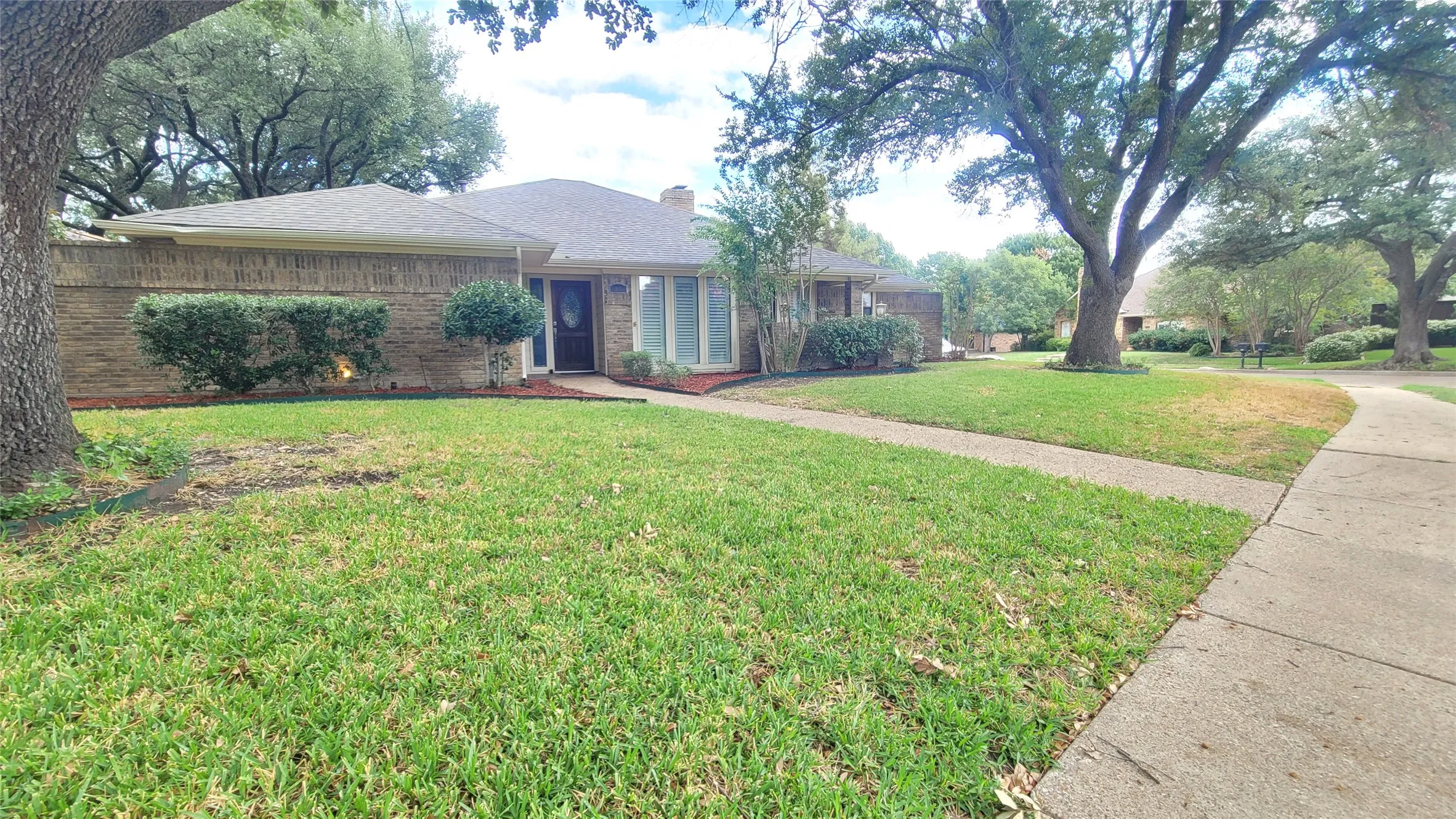 Single-story brick exterior featuring a multi-gabled roofline, concrete walkway, and expansive front lawn with mature trees