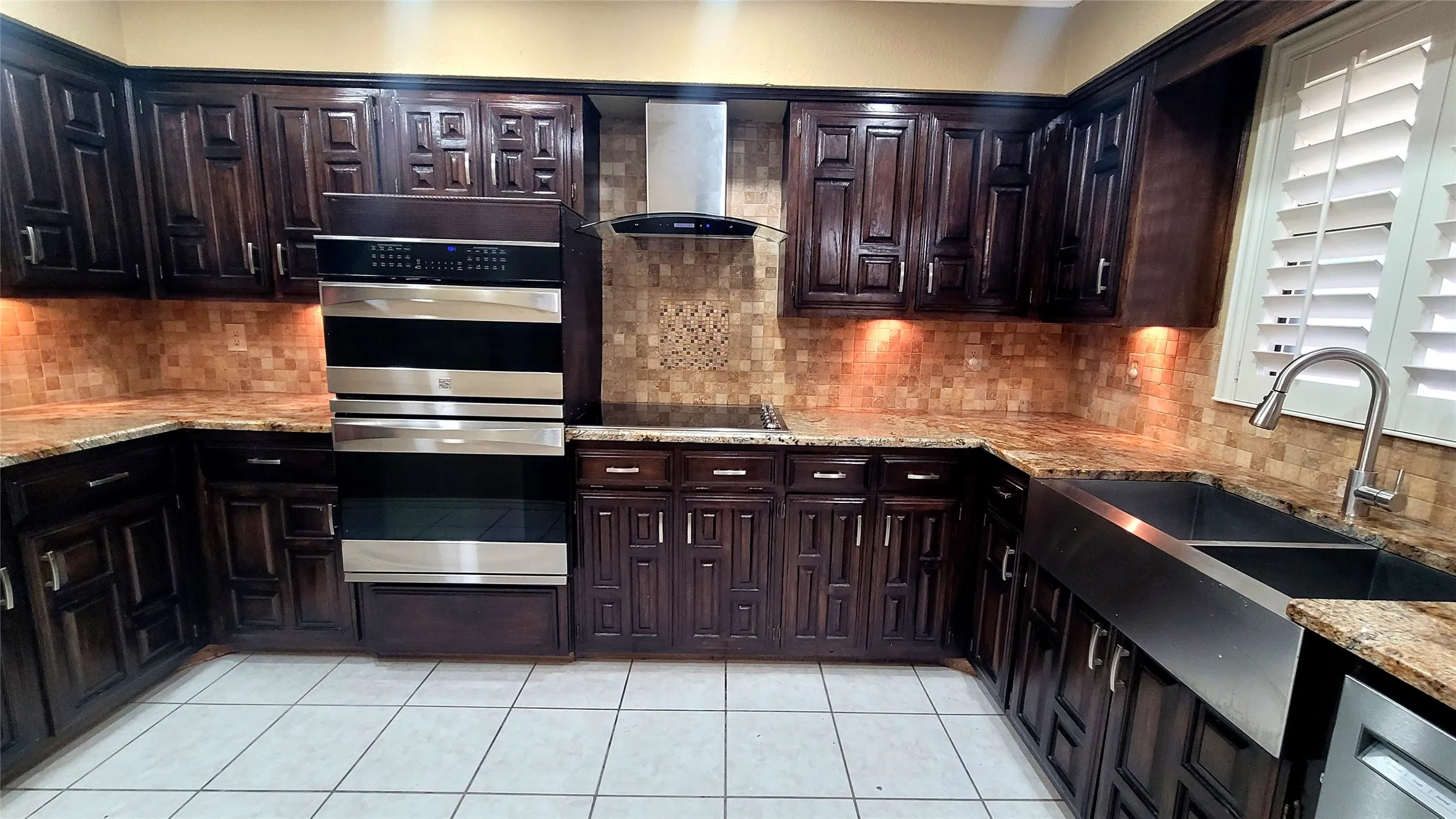 Kitchen featuring dark wood cabinetry, stainless steel double wall ovens, a farmhouse sink, stone countertops, and a mosaic tile backsplash