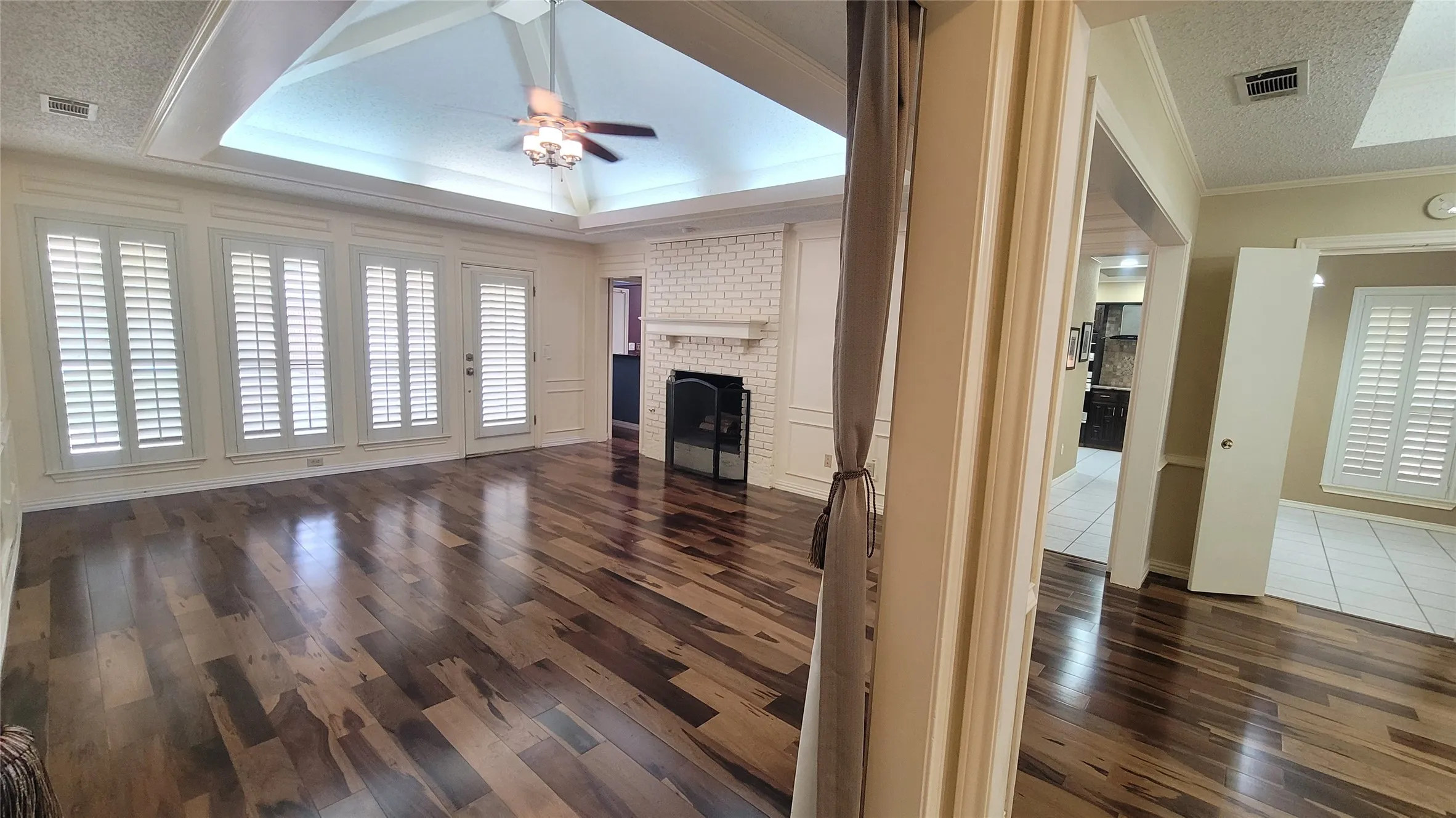 Living area featuring wood-finish flooring, tray ceiling with recessed lighting, whitewashed brick fireplace with mantel, and plantation shutters