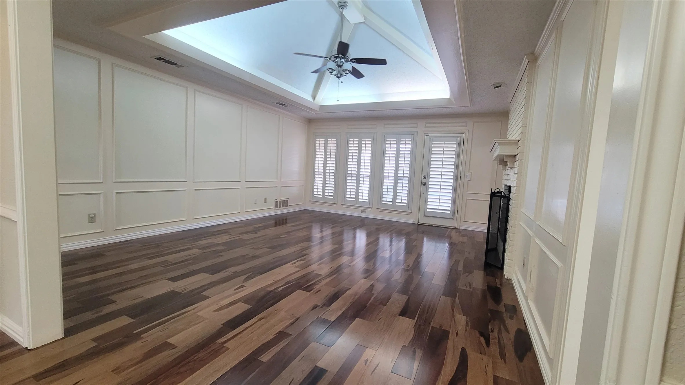 Spacious room showcasing rich wood-finish flooring, coffered ceiling with recessed lighting and ceiling fan, white paneled walls with wainscoting detail, and multiple glass doors with plantation shutters providing natural light