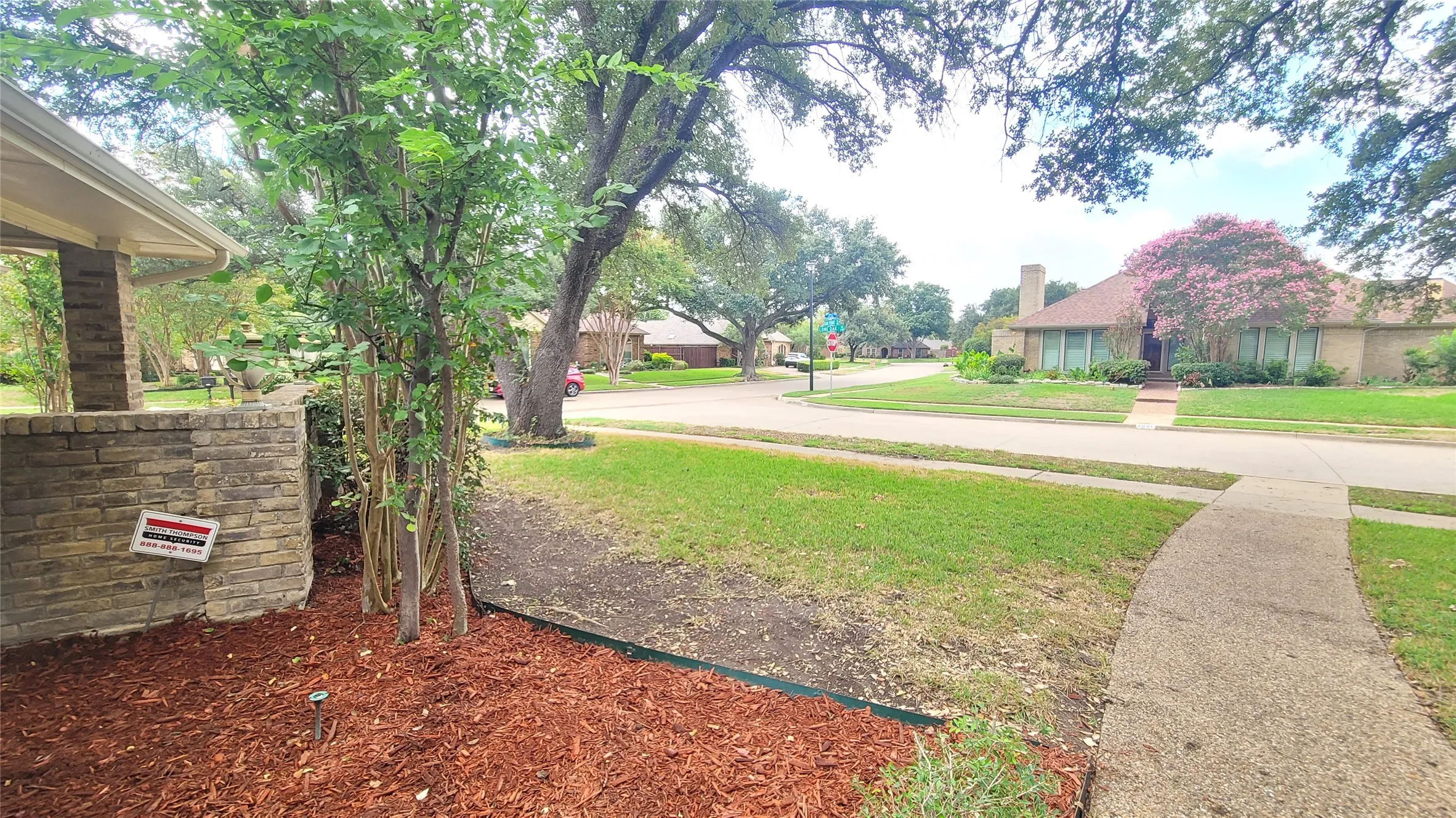 Covered front porch with brick column, landscaped garden beds, mature trees, concrete walkway, and lawn