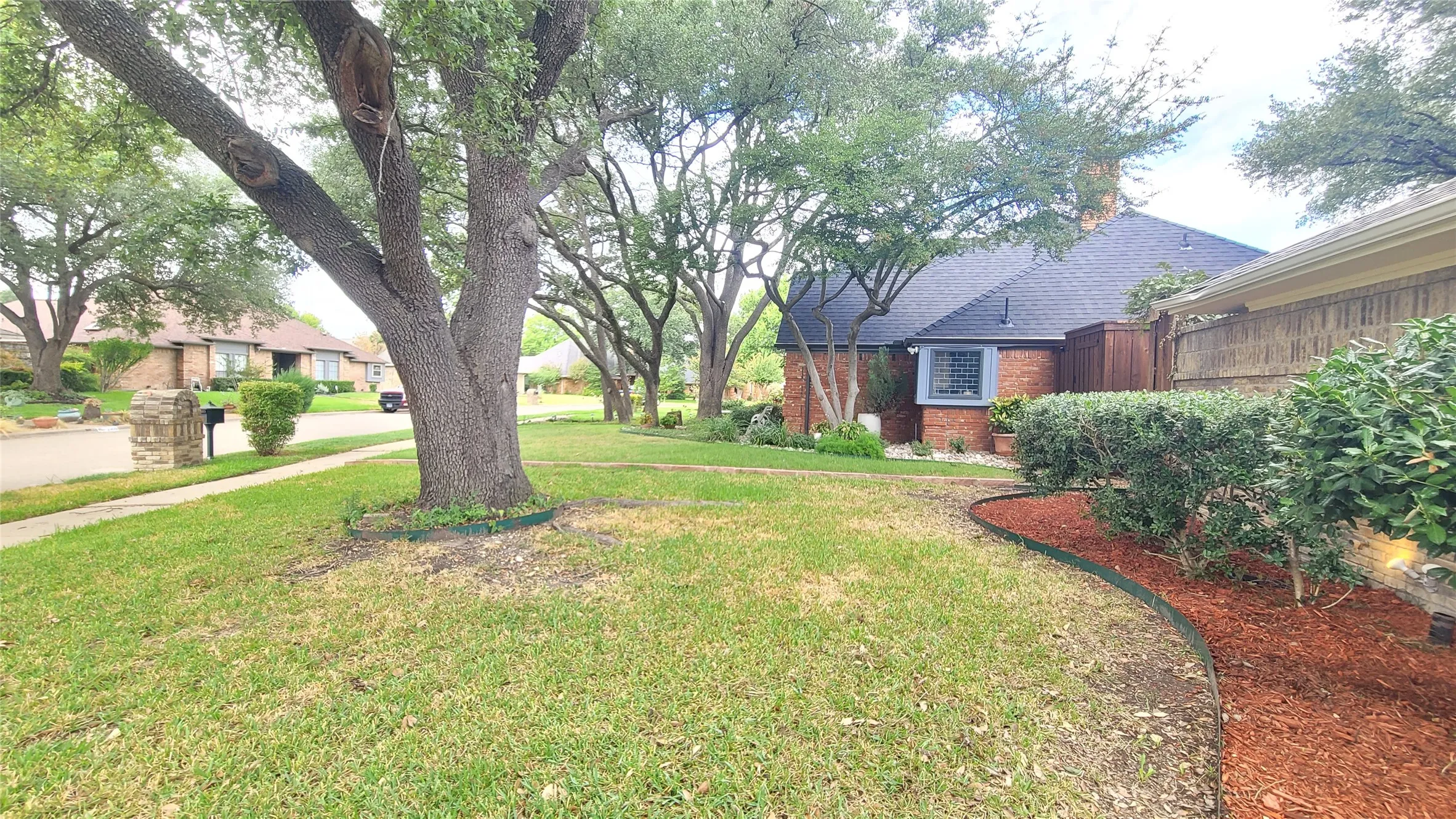 Front yard featuring mature shade trees, established landscaping with mulch beds, and a brick exterior facade