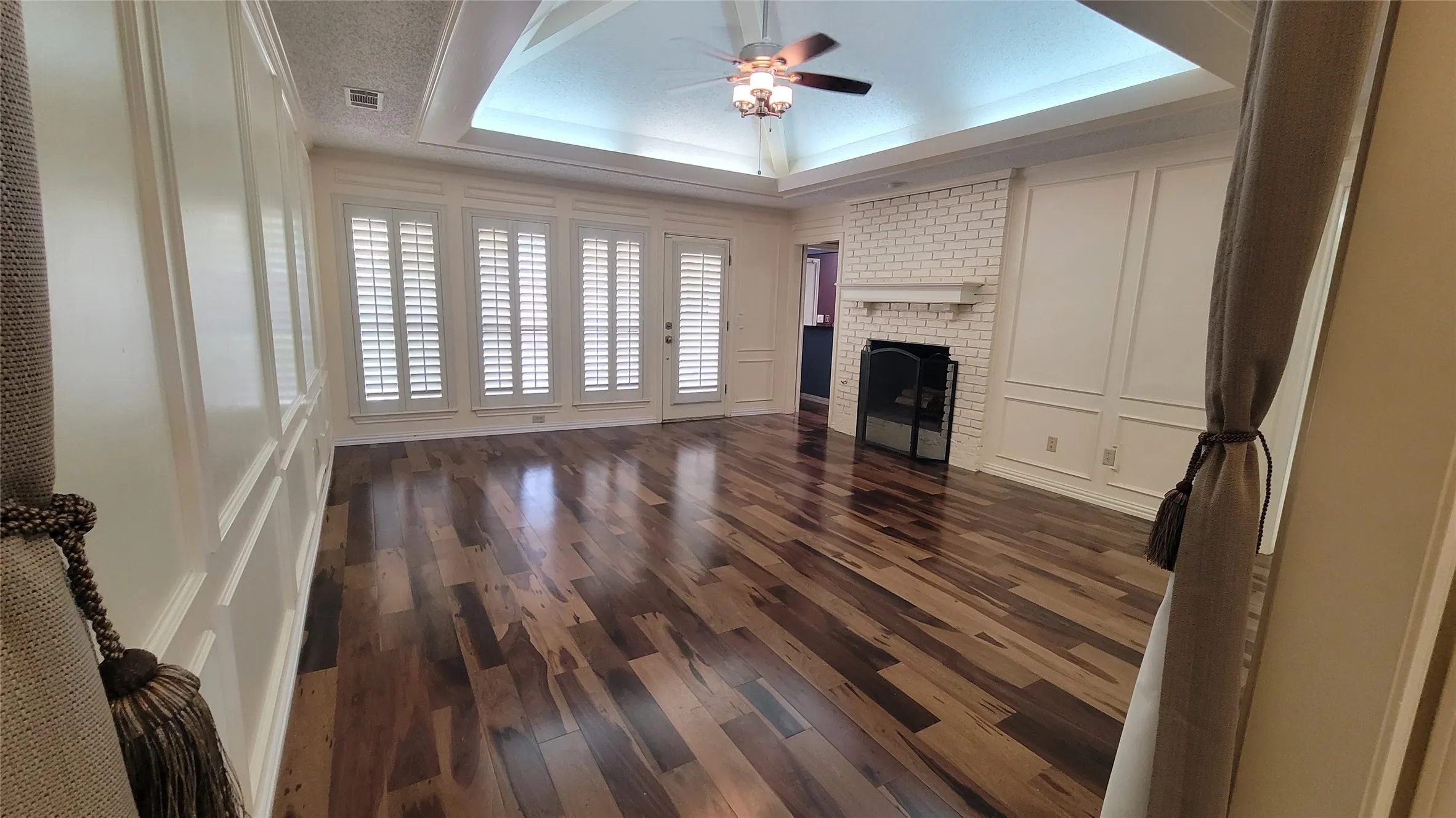 Tray ceiling with recessed lighting, wood-finish flooring, brick fireplace with a wood mantel, and walls featuring wainscoting and plantation shutters