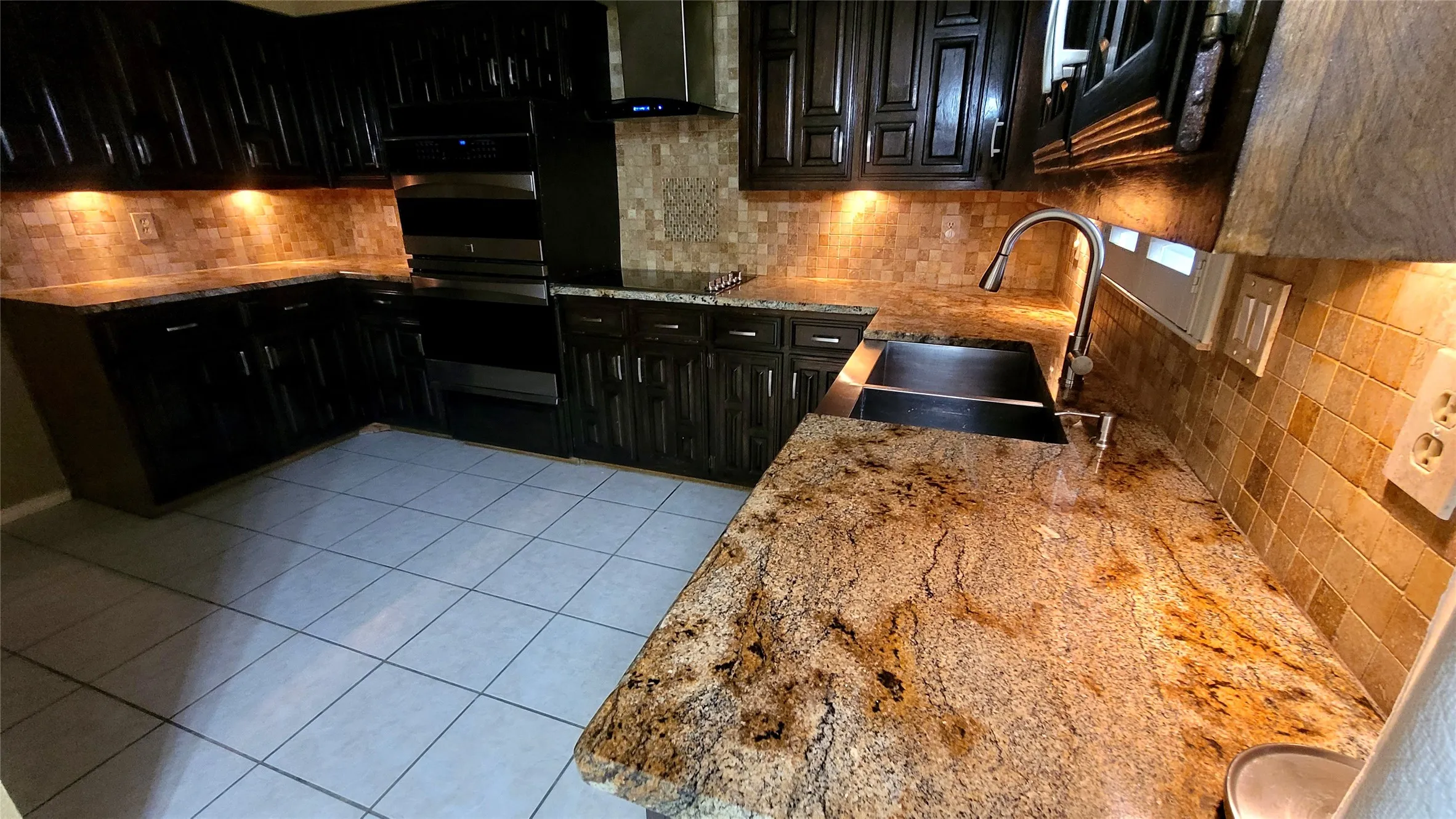Elegant kitchen featuring granite countertops, undermount sink with bronze faucet, custom dark cabinetry with under-cabinet lighting, ceramic tile flooring, and copper tile backsplash