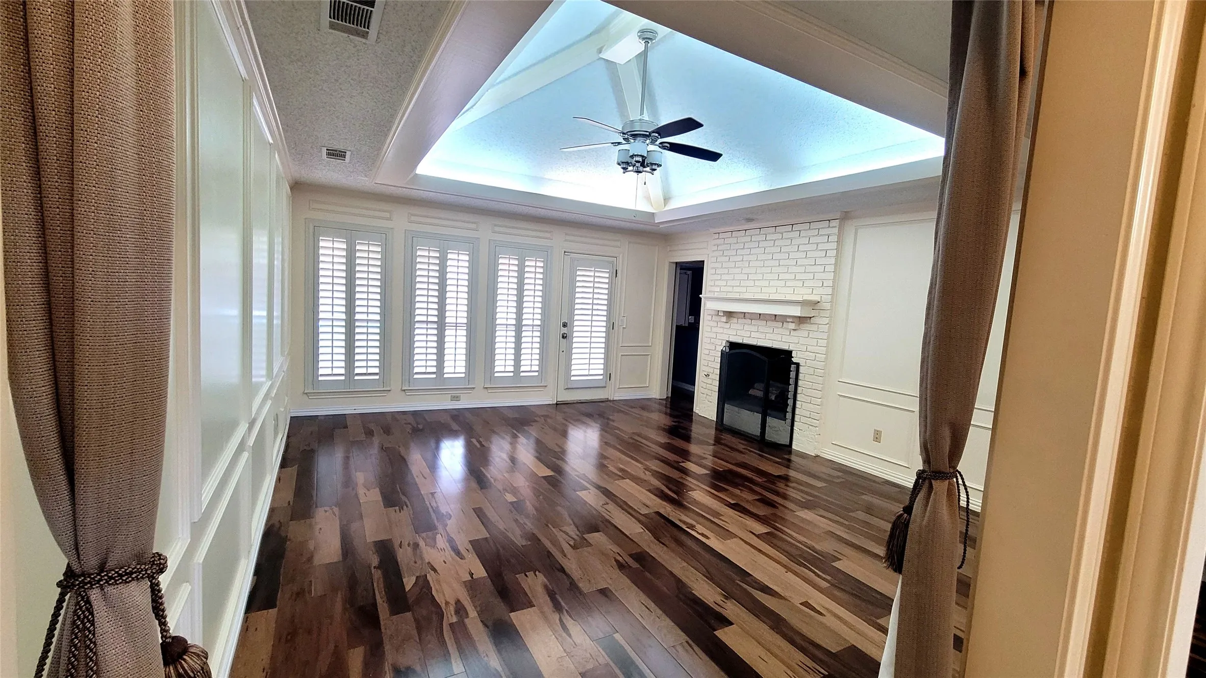 Vaulted tray ceiling with recessed lighting, wood-finish flooring, floor-to-ceiling wainscoting, white painted brick fireplace with mantle, and plantation shutters