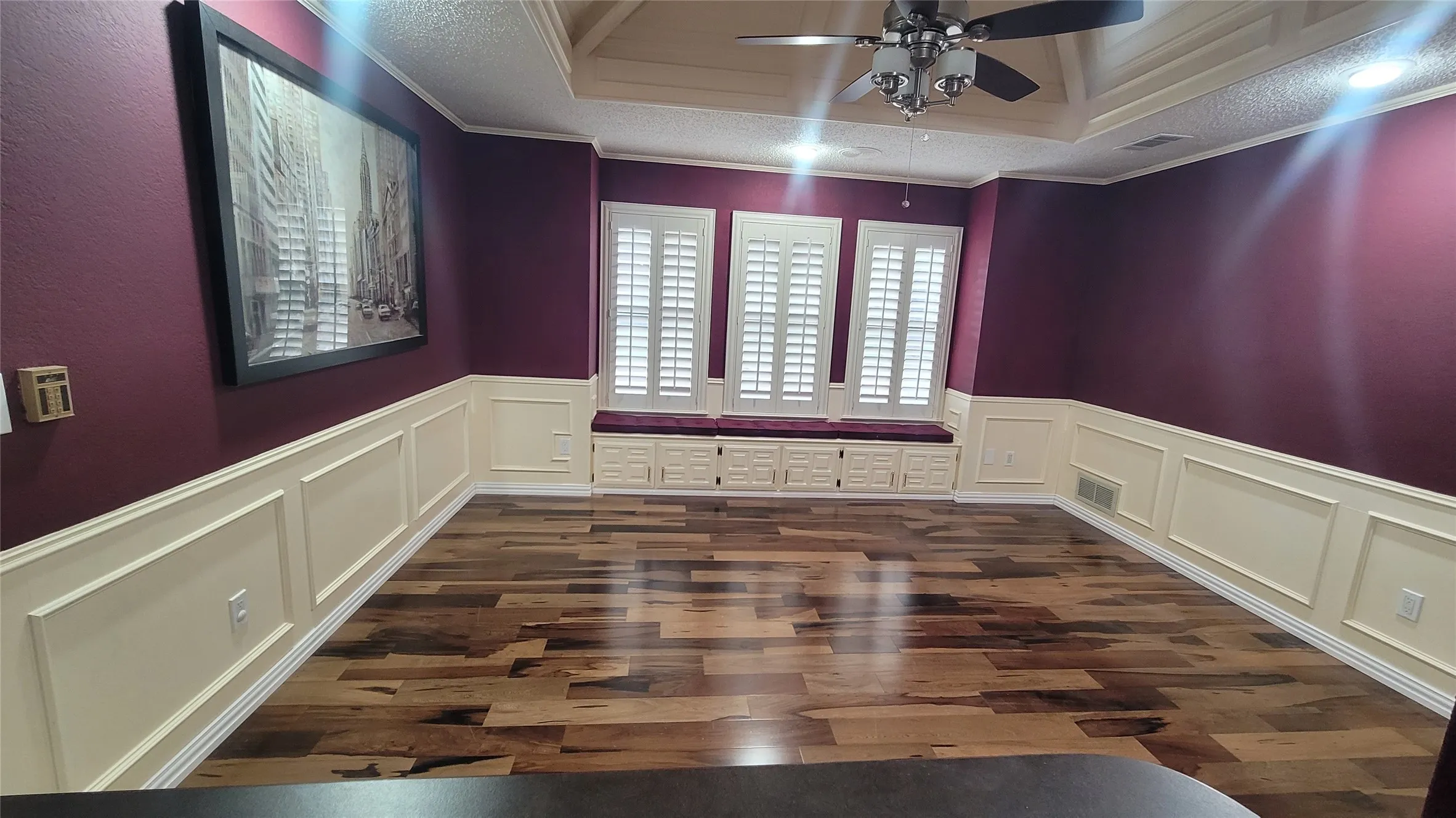 Tray ceiling with a central ceiling fan, wood-finish flooring, custom wall molding, and built-in window seating with shuttered windows