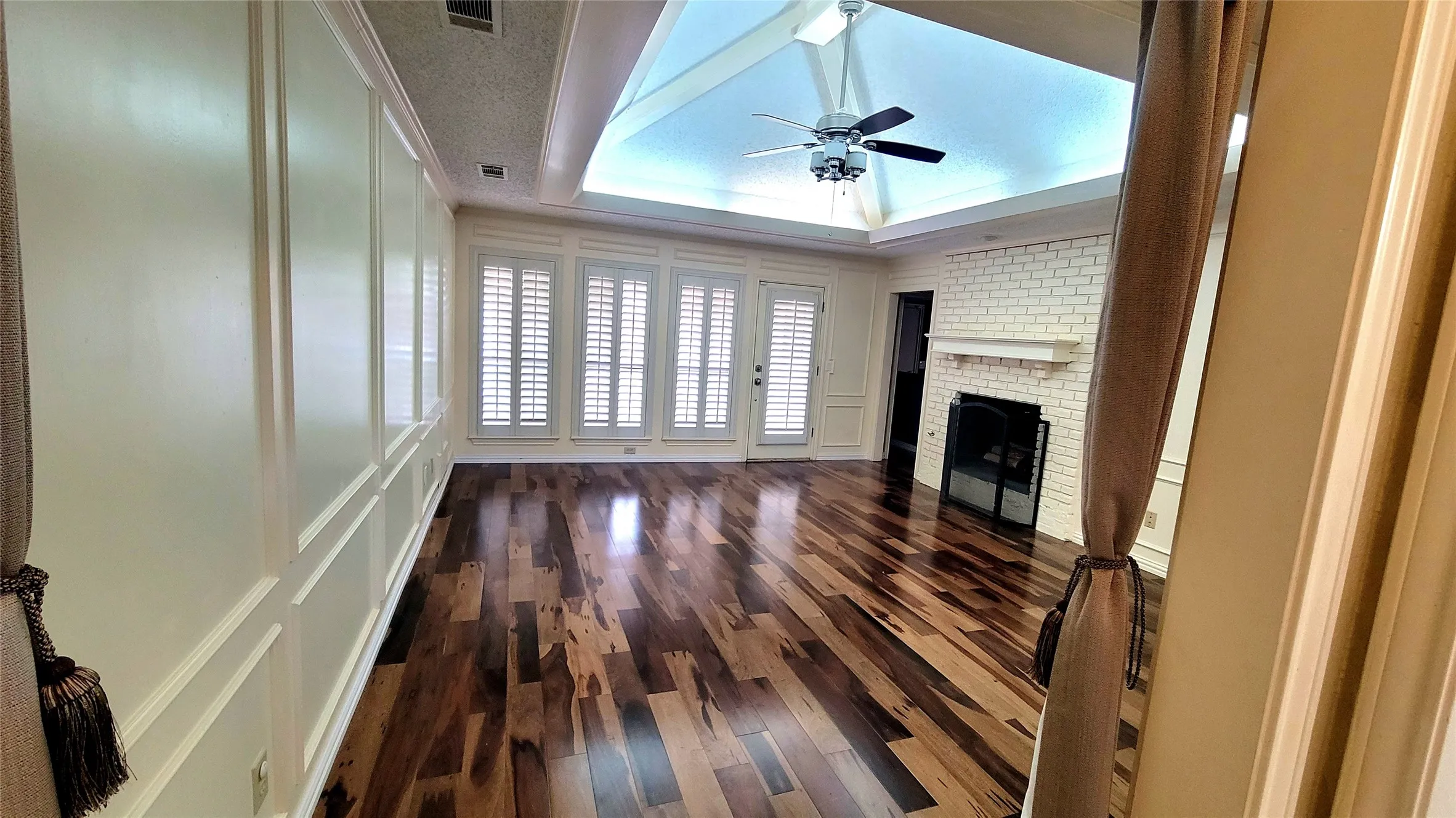 Vaulted ceiling featuring a central light fixture, wood-finish flooring, brick fireplace with a mantel, wall paneling, and shuttered windows