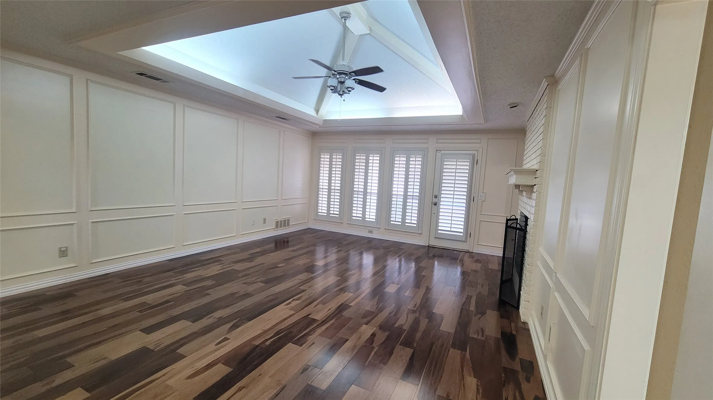 Vaulted ceiling with a central fan, wood-finish flooring, wainscoting wall paneling, brick fireplace with a mantel, and plantation shutters
