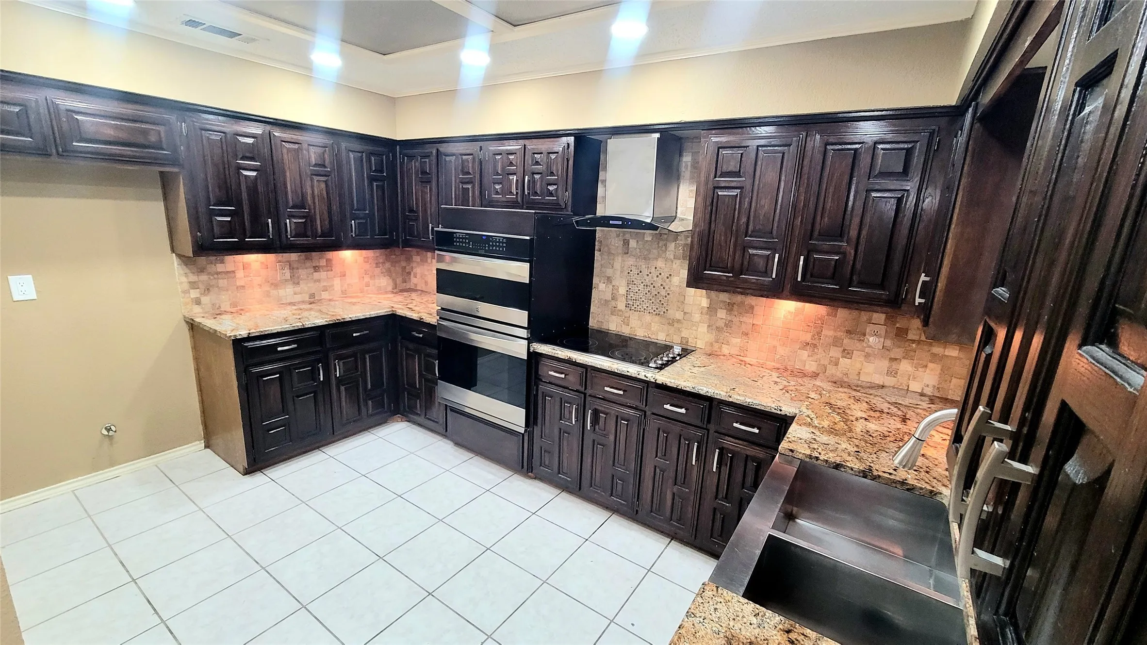 Kitchen featuring dark wood cabinetry, patterned stone countertops, stainless steel double wall oven, stainless steel range hood, and tile flooring