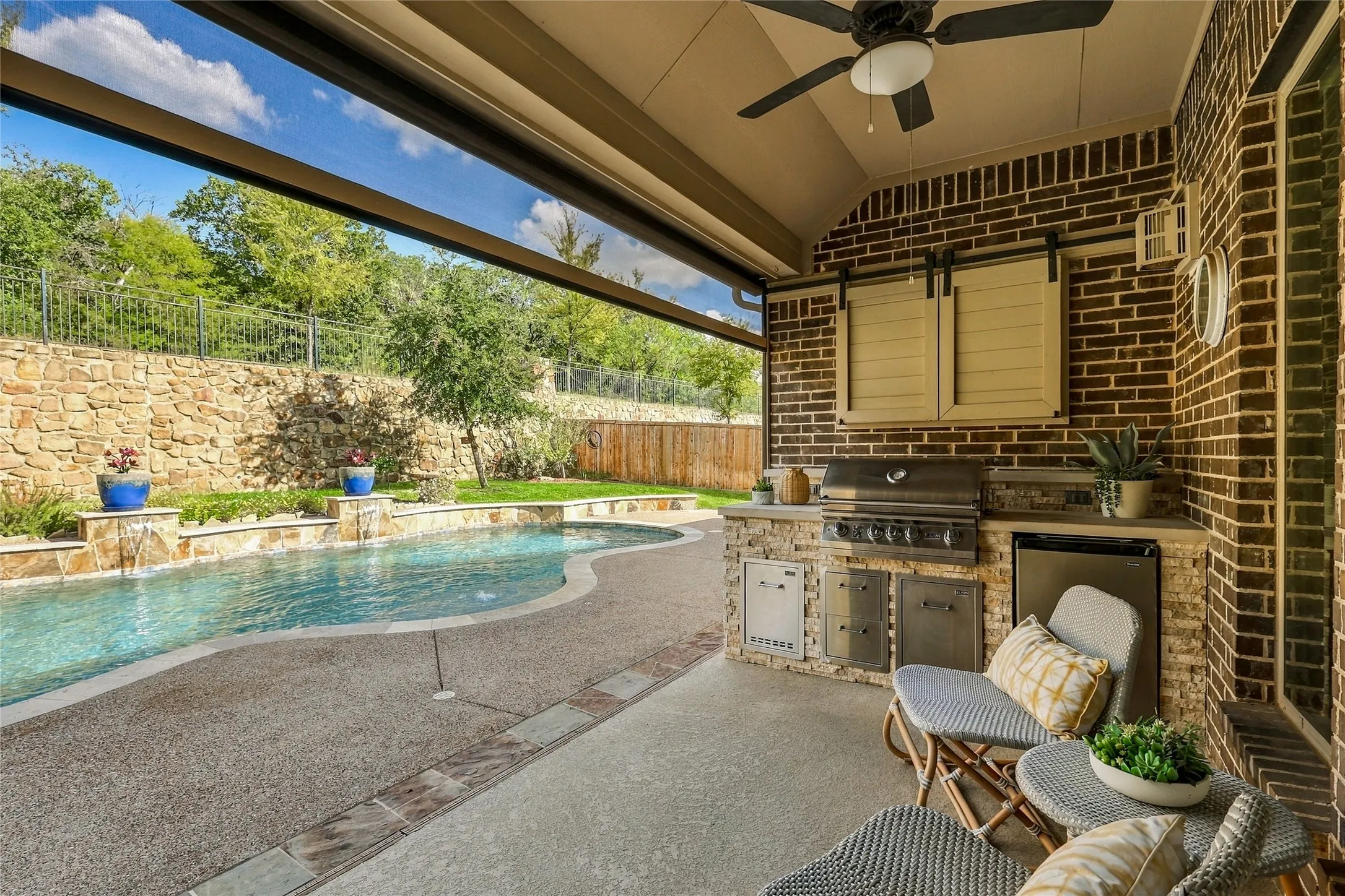 Covered patio featuring an outdoor kitchen with stainless steel appliances, stone masonry, a built-in ceiling fan, and a swimming pool with a stone retaining wall
