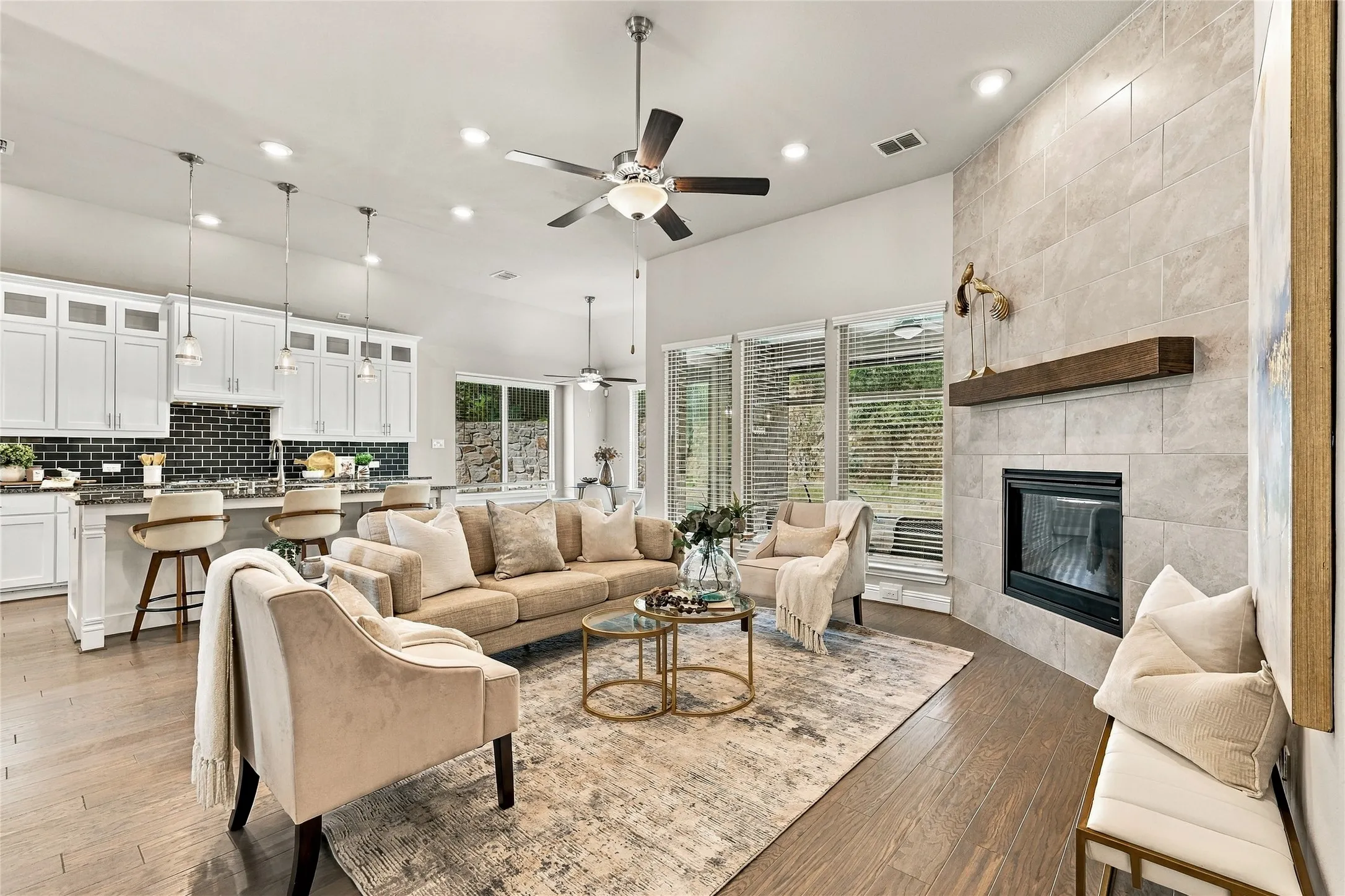 Open-concept interior featuring wood-finish flooring, a full-height stone-surround fireplace with a wooden mantel, white cabinetry, a kitchen island with pendant lighting, and recessed ceiling lights