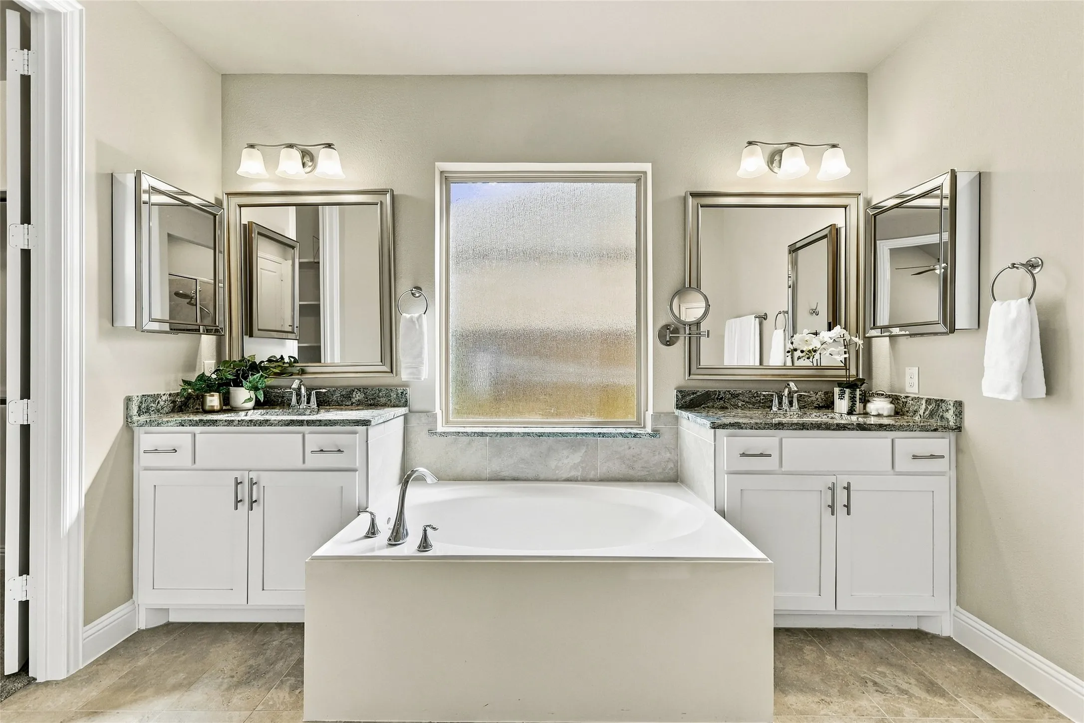 Dual white vanities featuring stone-finish countertops, undermount sinks, framed mirrors, and central soaking tub with an arched faucet and frosted glass window