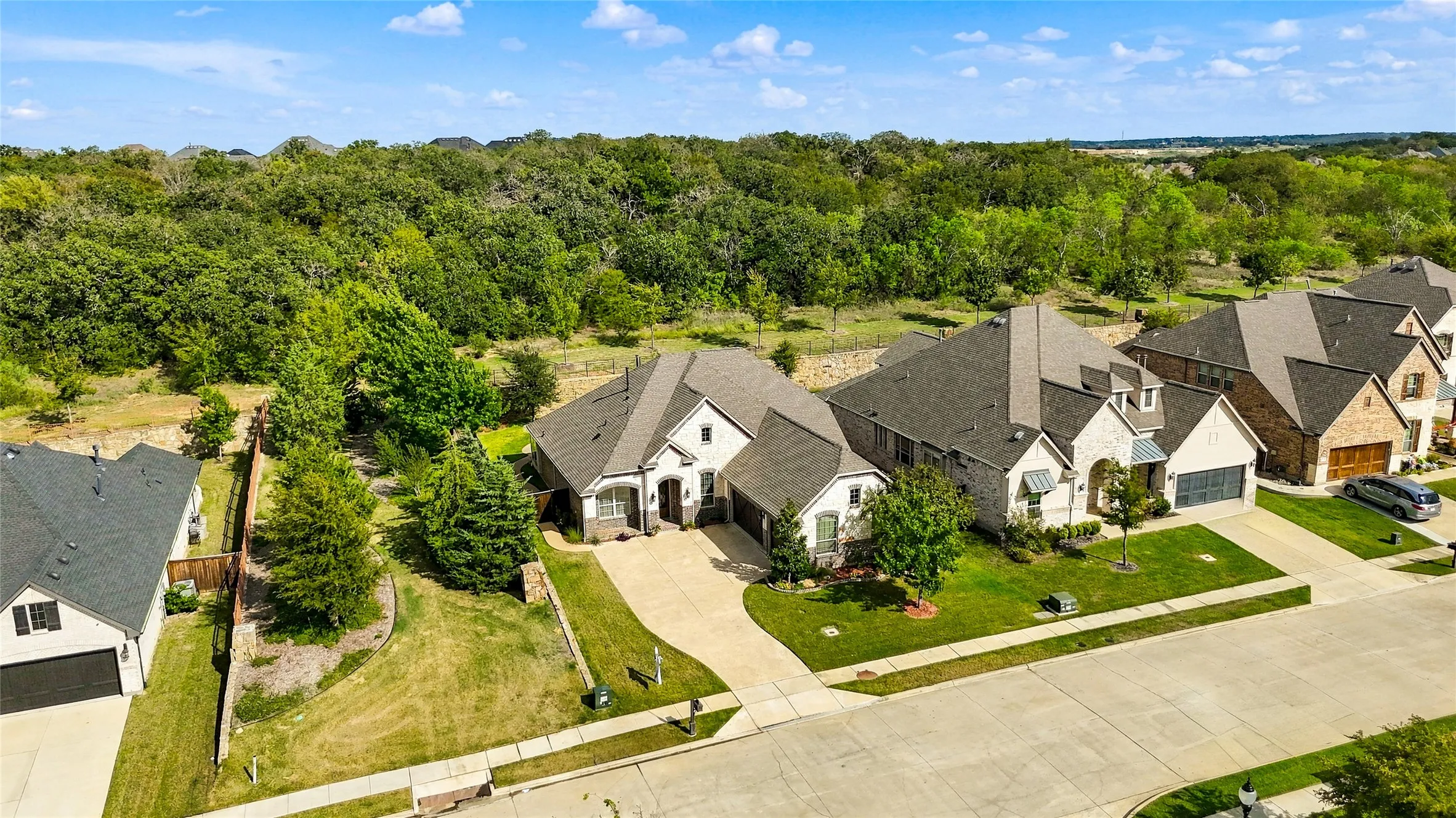 Aerial perspective featuring a multi-gabled brick and masonry exterior, asphalt shingle roofing, concrete driveway, and a perimeter fence line bordering an expanse of mature trees