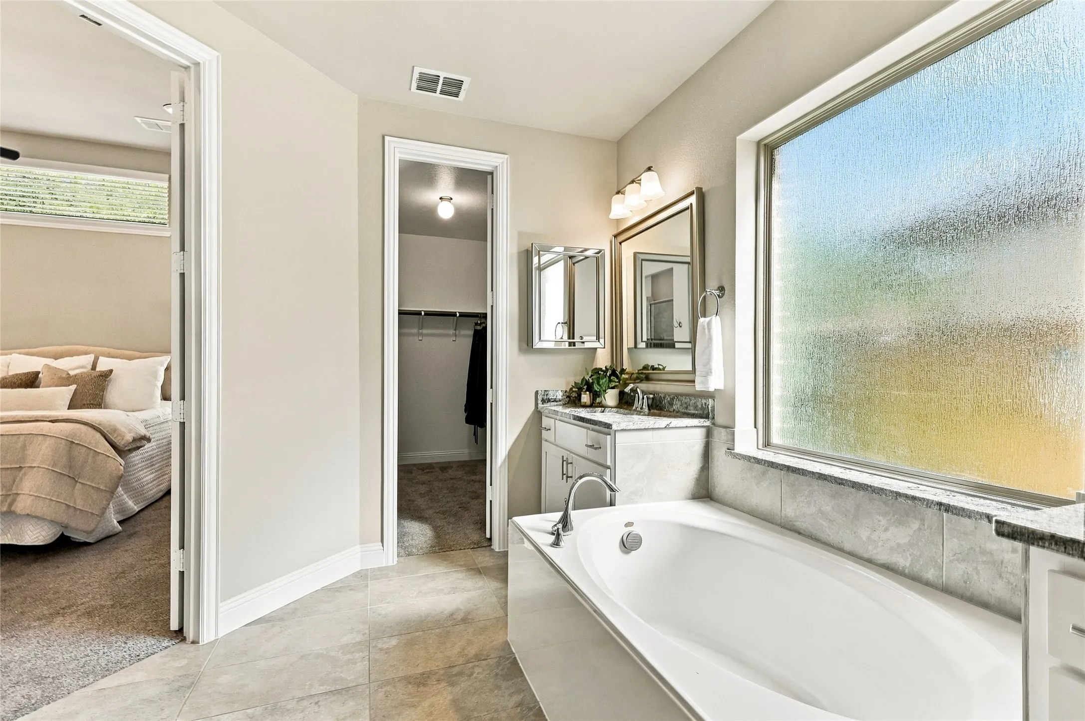 Bathroom featuring a deep soaking tub with tile surround, large privacy-glass window, vanity with stone-style countertop, and wall-mounted light fixtures