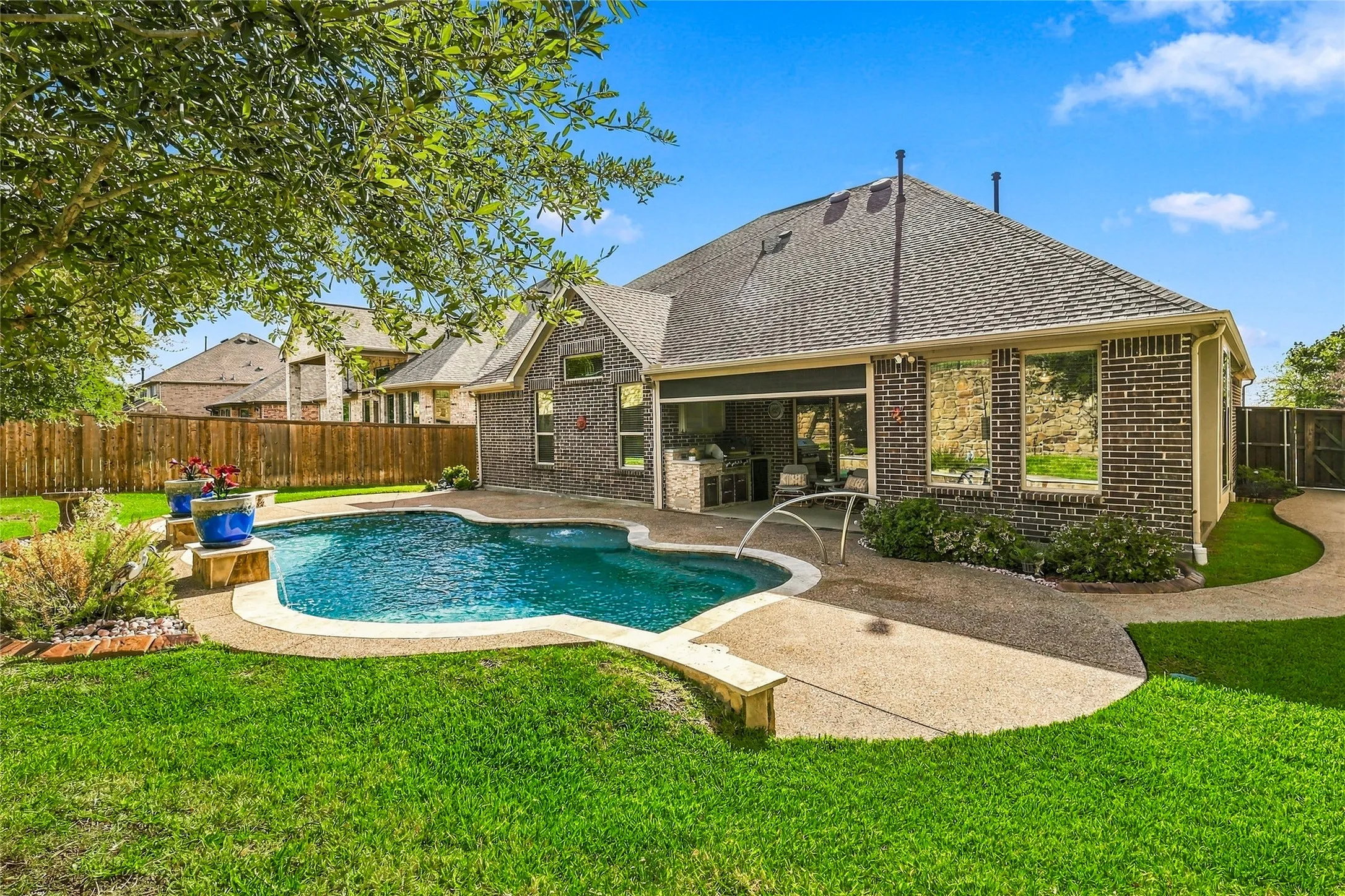 Brick exterior residence featuring an in-ground pool, covered patio with outdoor kitchen, concrete decking, and fenced backyard