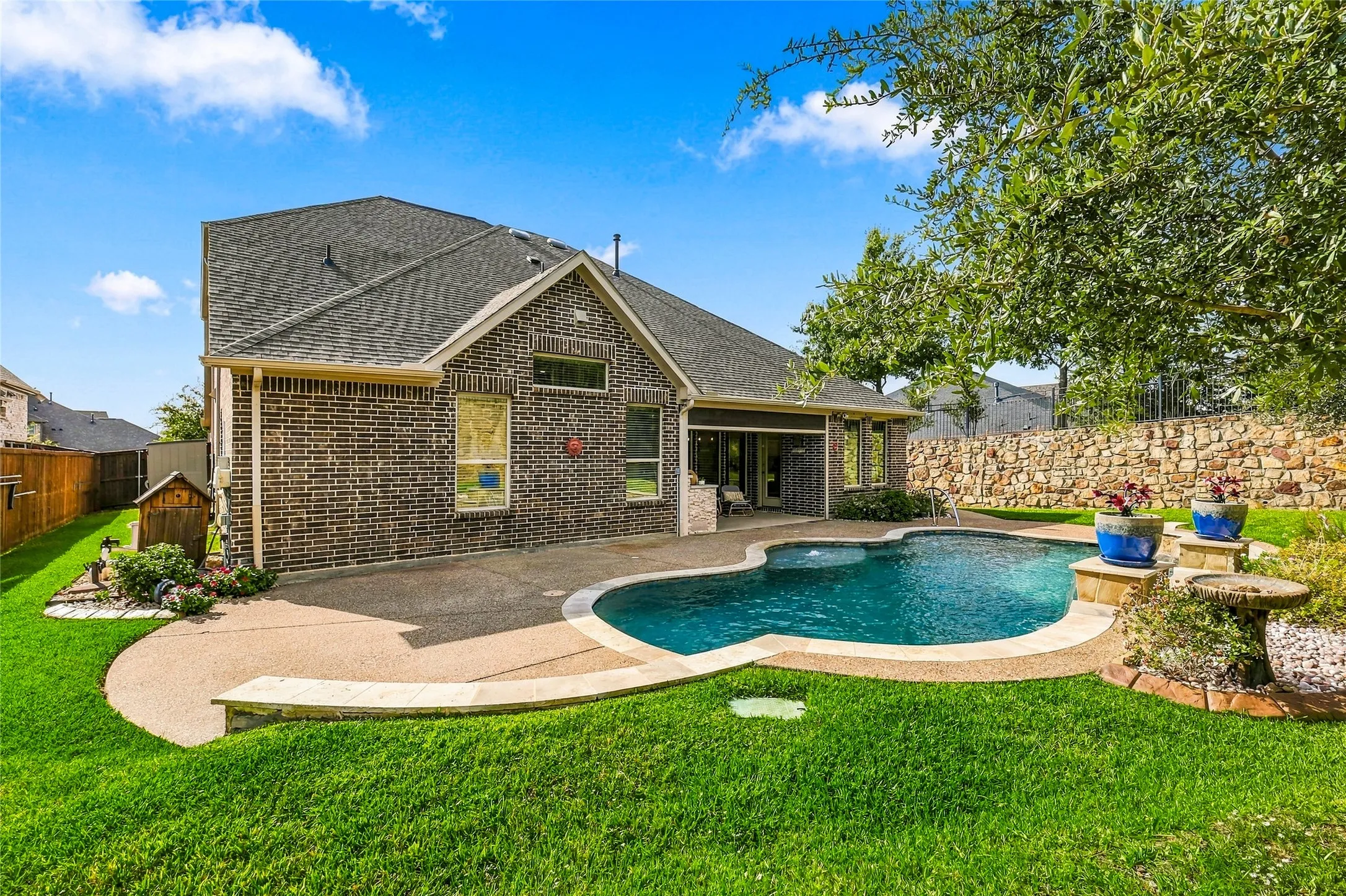 Brick exterior home featuring a rear swimming pool, concrete patio, stone retaining wall, and manicured lawn