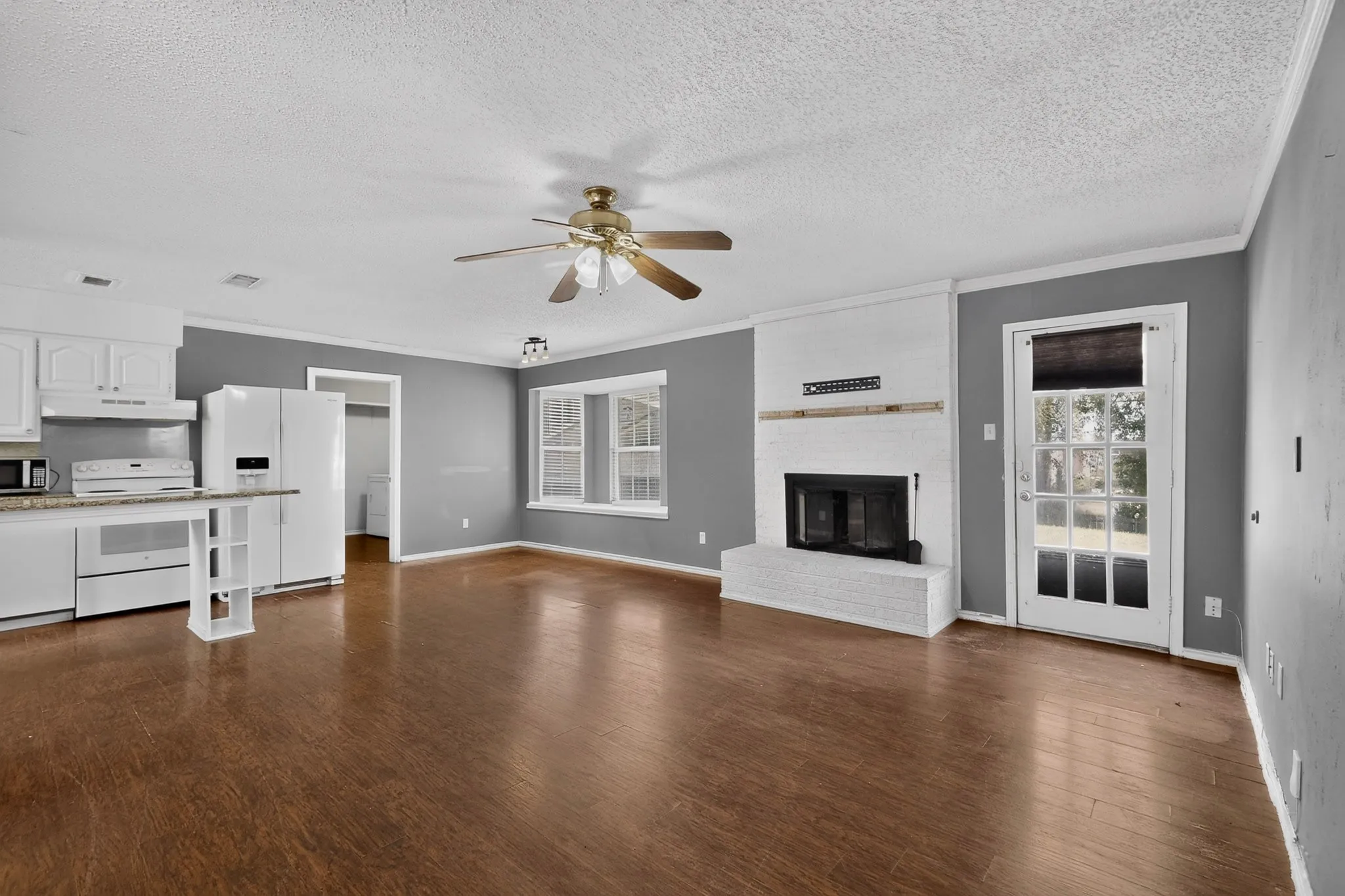 Wood-finish flooring, white painted brick fireplace with a mantle, built-in kitchen cabinetry, ceiling fan, and multi-light glass panel exterior door