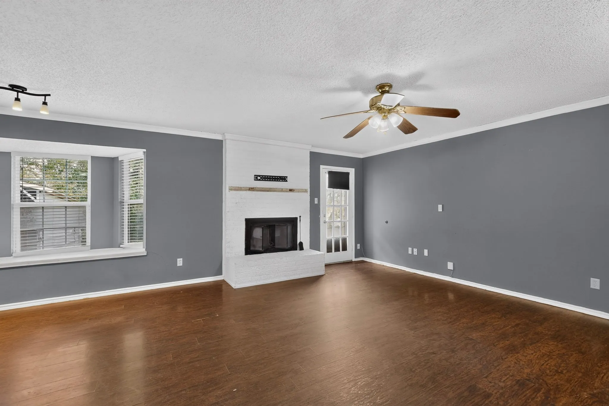 Living area featuring wood-finish flooring, a painted brick fireplace with a wood mantel, a multi-paneled bay window, and a ceiling fan with integrated lighting