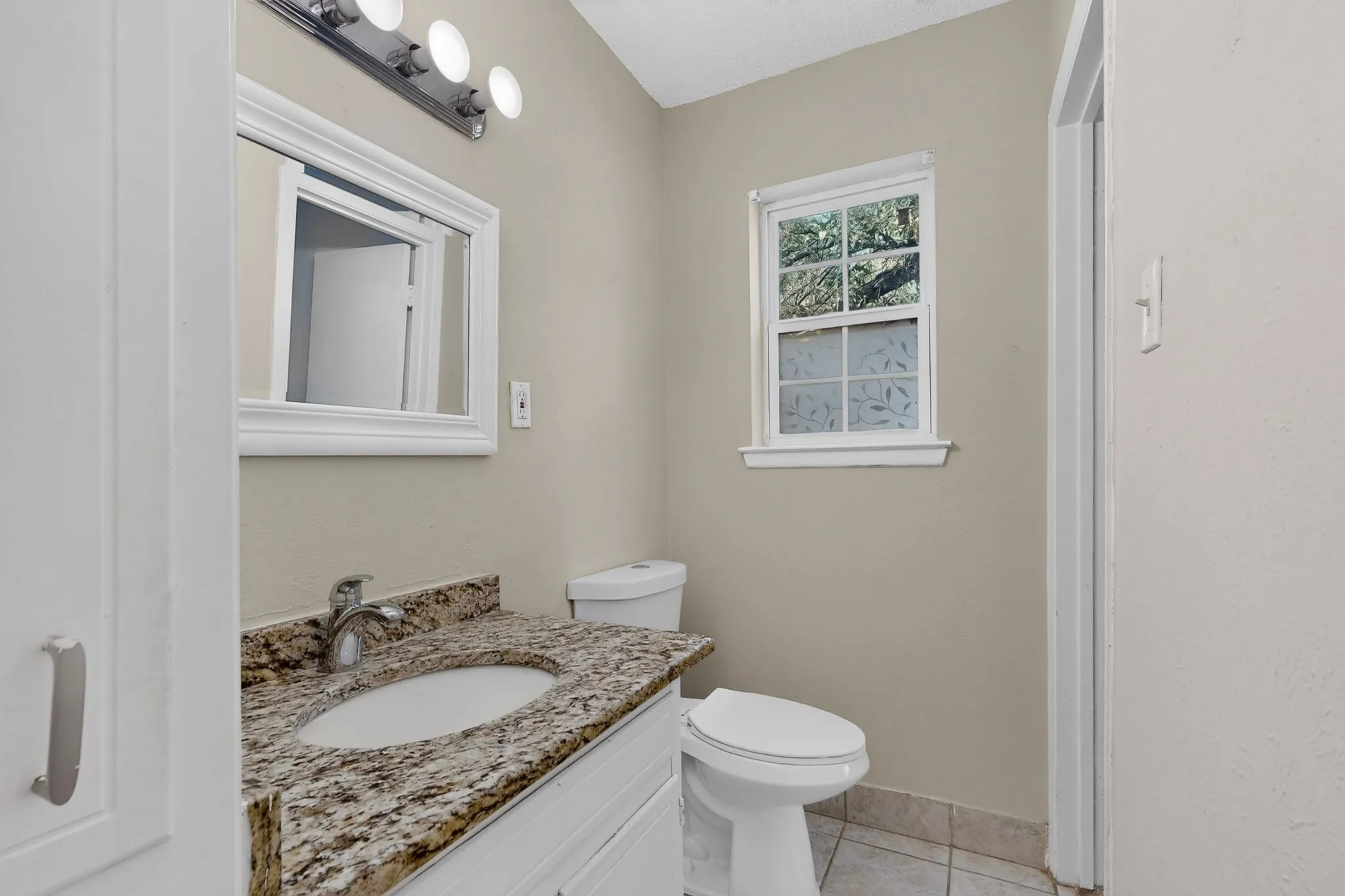 Stone-topped vanity with an integrated oval sink, three-light wall-mounted fixture, framed vanity mirror, and ceramic tile flooring