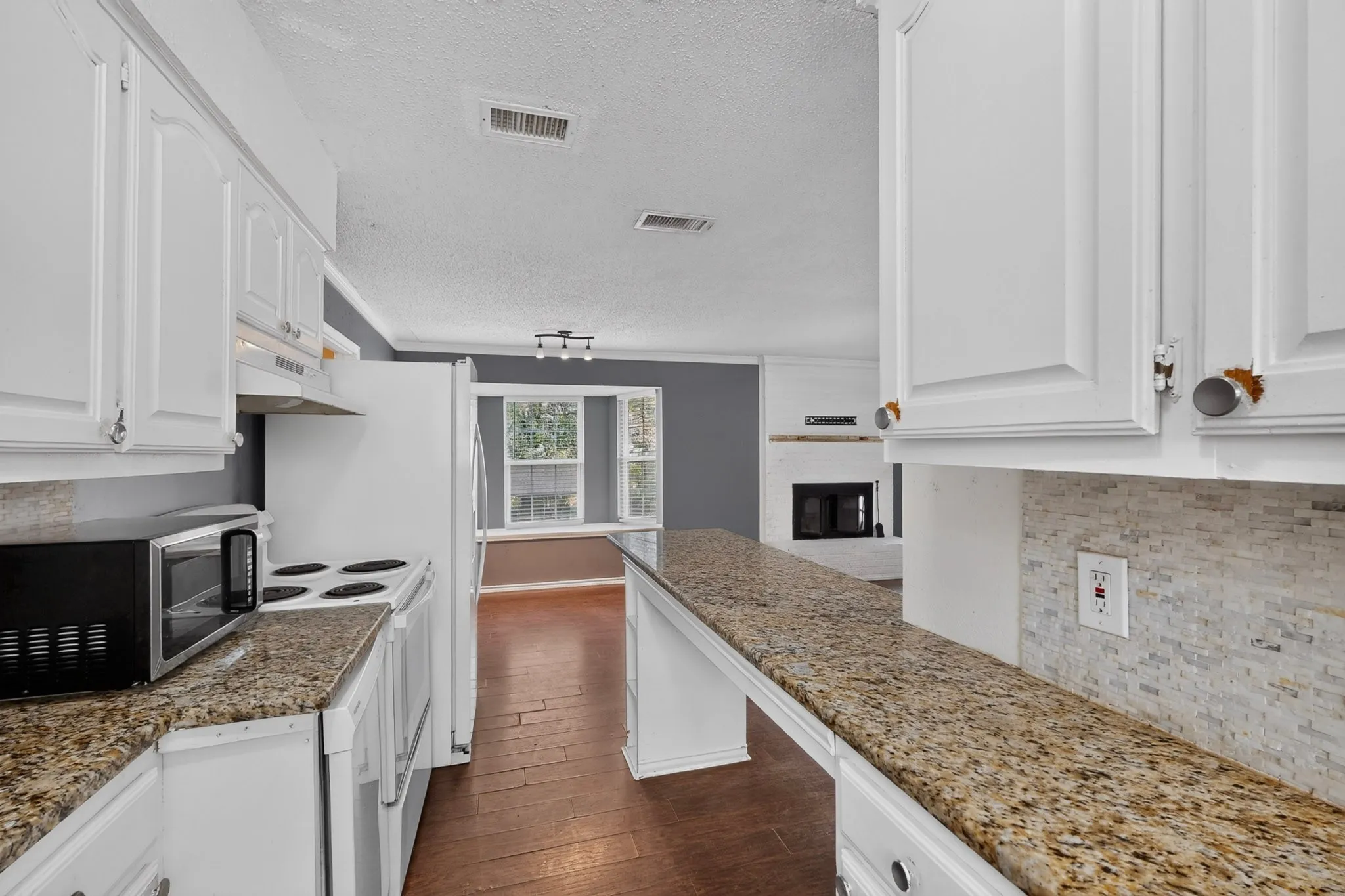 Kitchen featuring white cabinetry, stone-finish countertops, tile backsplash, and wood-finish flooring, extending toward a fireplace with a mantel