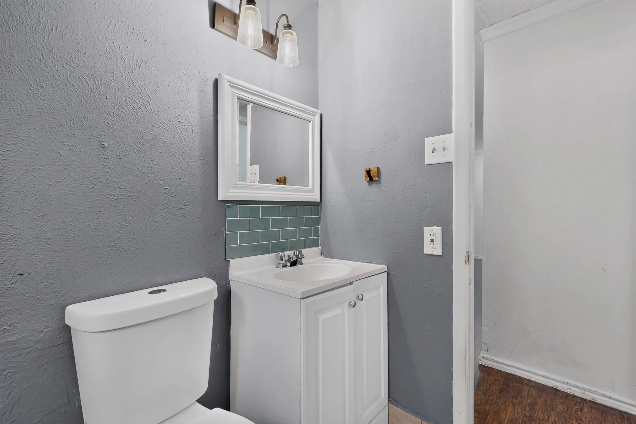 White vanity with integrated sink, teal subway tile backsplash, wall-mounted light fixture, framed mirror, and porcelain toilet