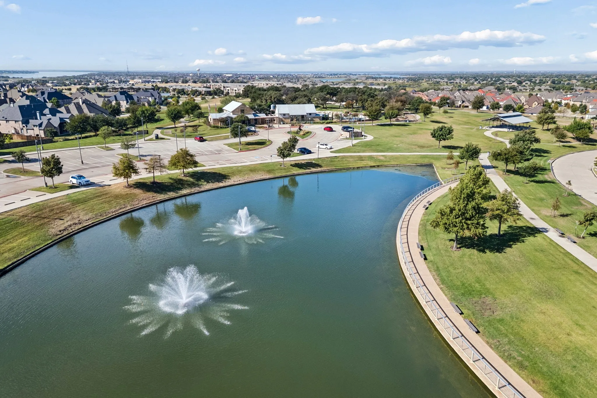 Pond featuring dual aerating fountains, concrete paved perimeter walkway, surrounding manicured green space, and mature trees