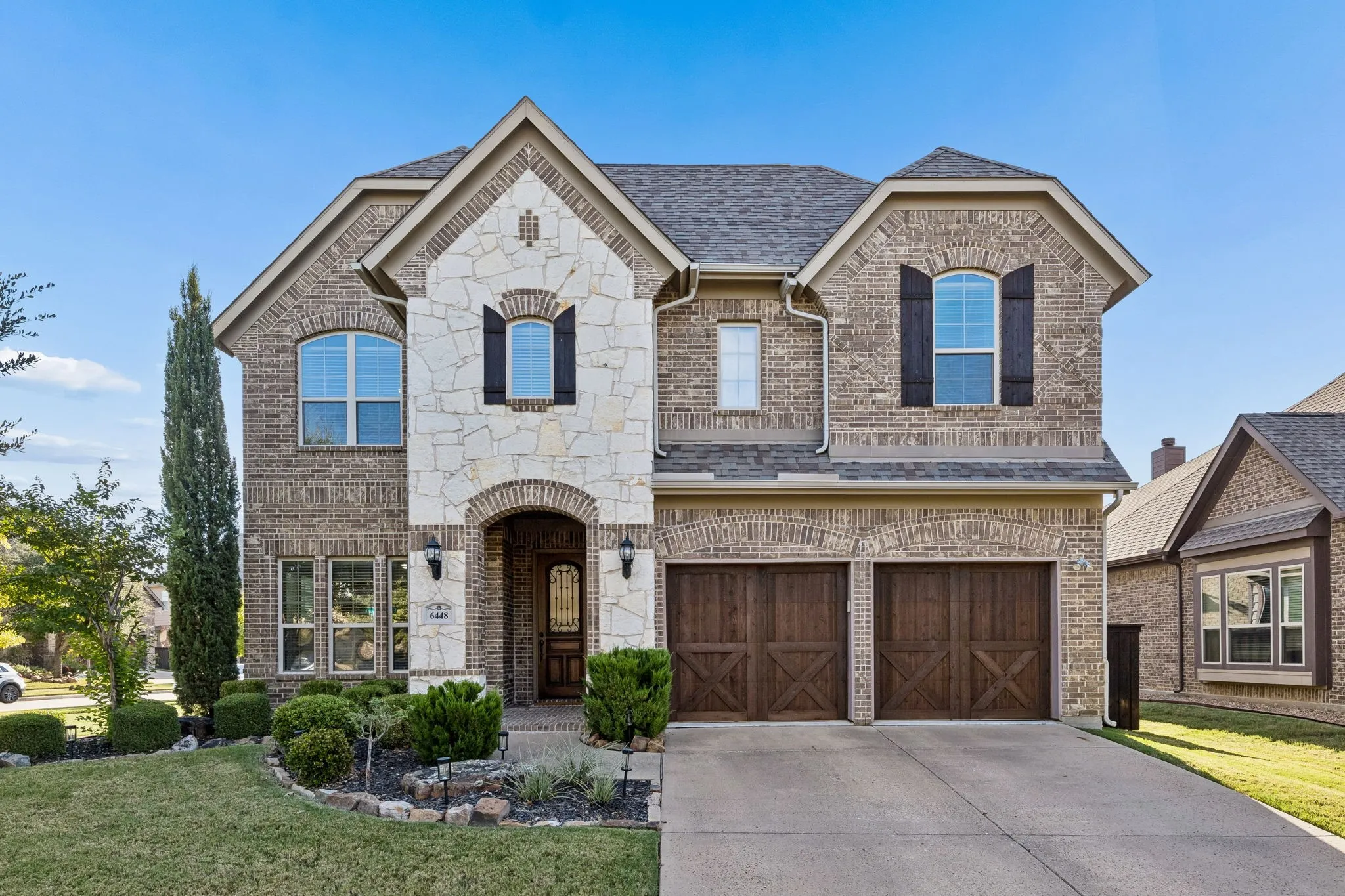 Two-story brick and stone facade with arched entryway, decorative wood-finish shutters, double carriage-style garage doors, and arched window detailing