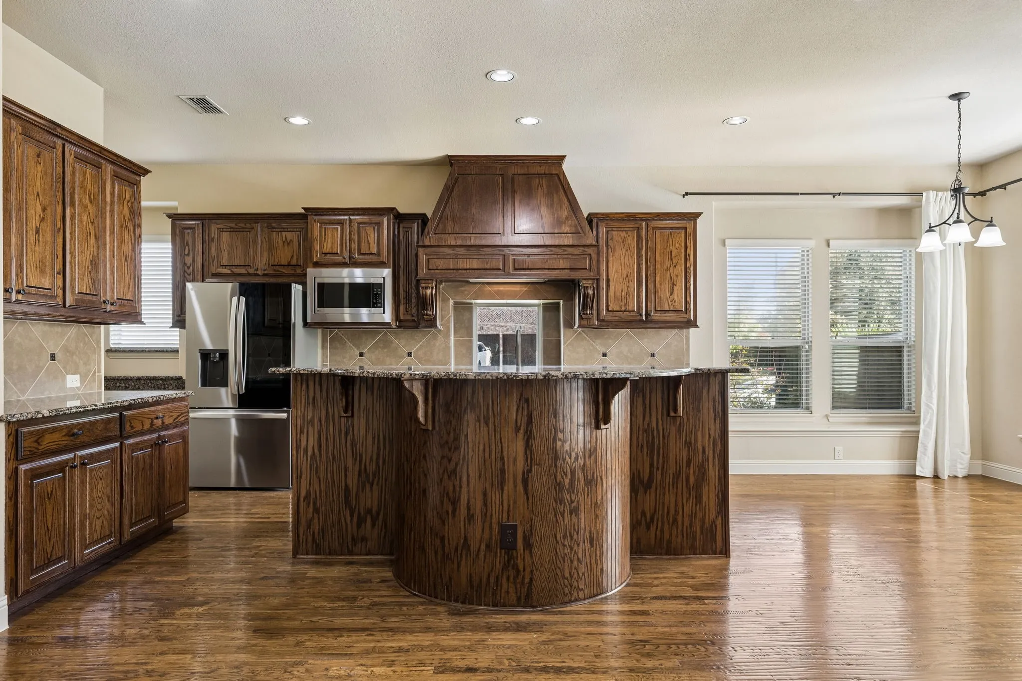 Kitchen featuring custom wood cabinetry, a curved island with stone countertops, stone tile backsplash, stainless steel appliances, and wood-finish flooring